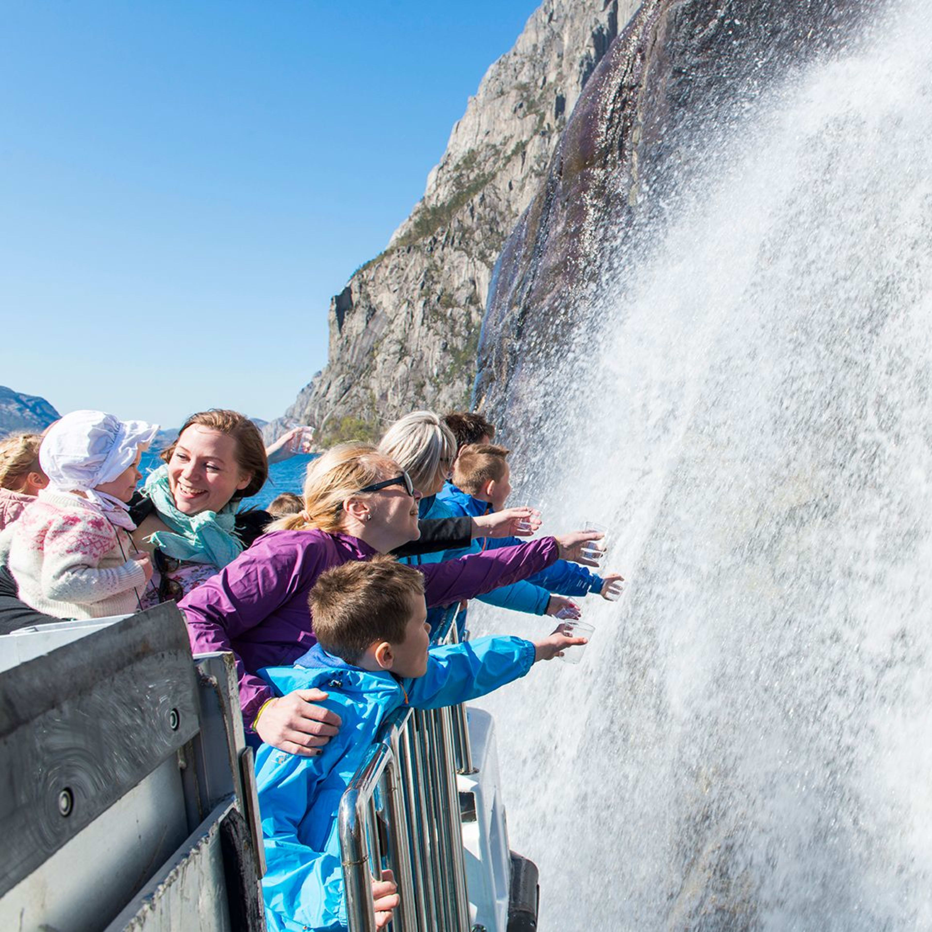 Turister på fjordcruise på Lysefjorden strekker seg mot Hengjanefossen under en knallblå himmel.