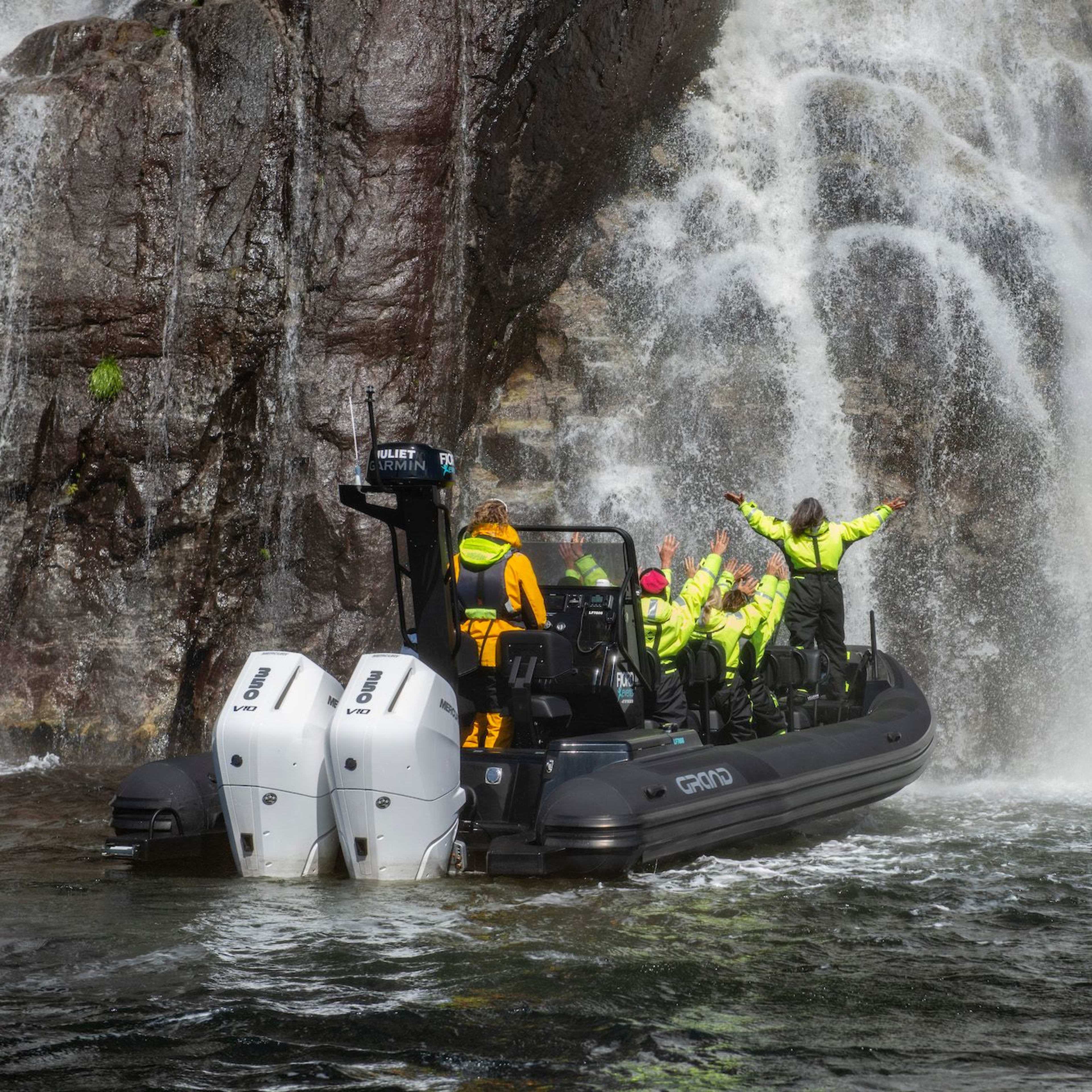 Ein schwarzes Festrumpfschlauchboot liegt in der Nähe eines Wasserfalls im Lysefjord. Die Passagiere heben aufgeregt die Arme.