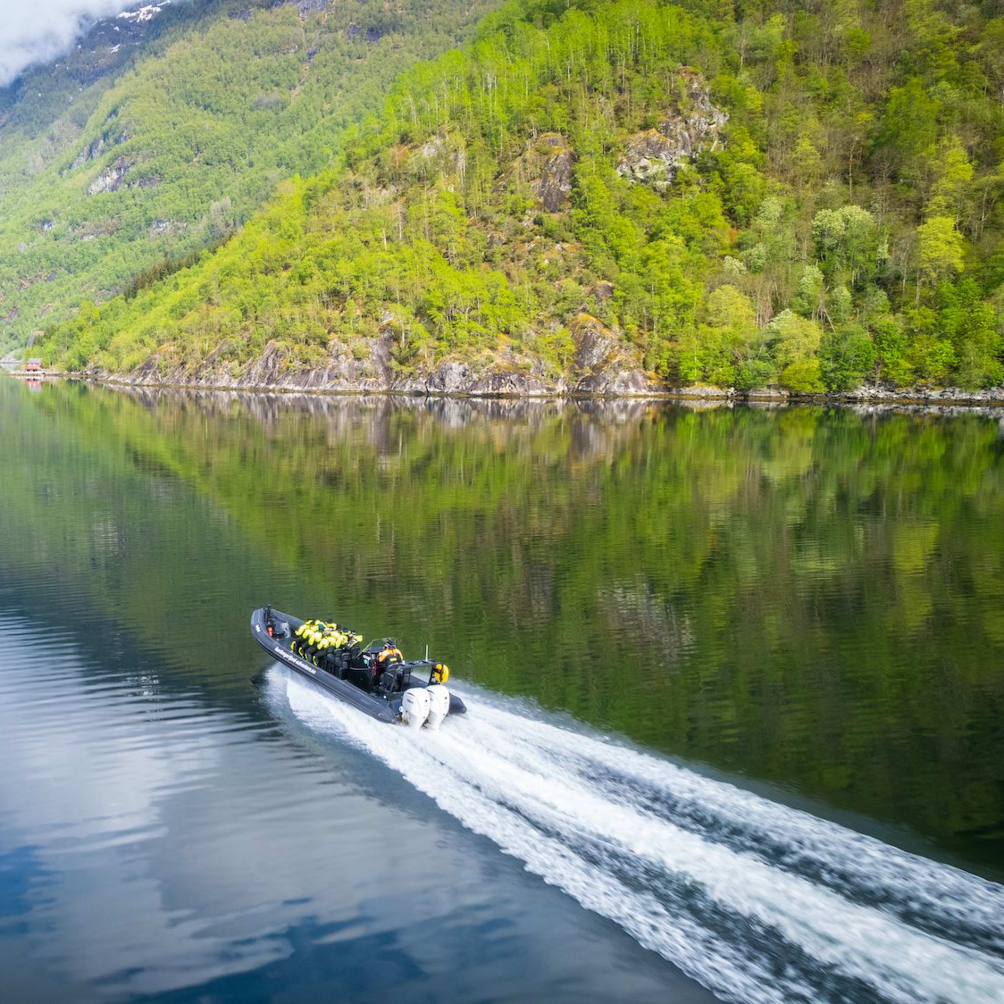 Boat with yellow-jacketed passengers speeds on reflective fjord, mountains and cloudy sky mirrored in water.