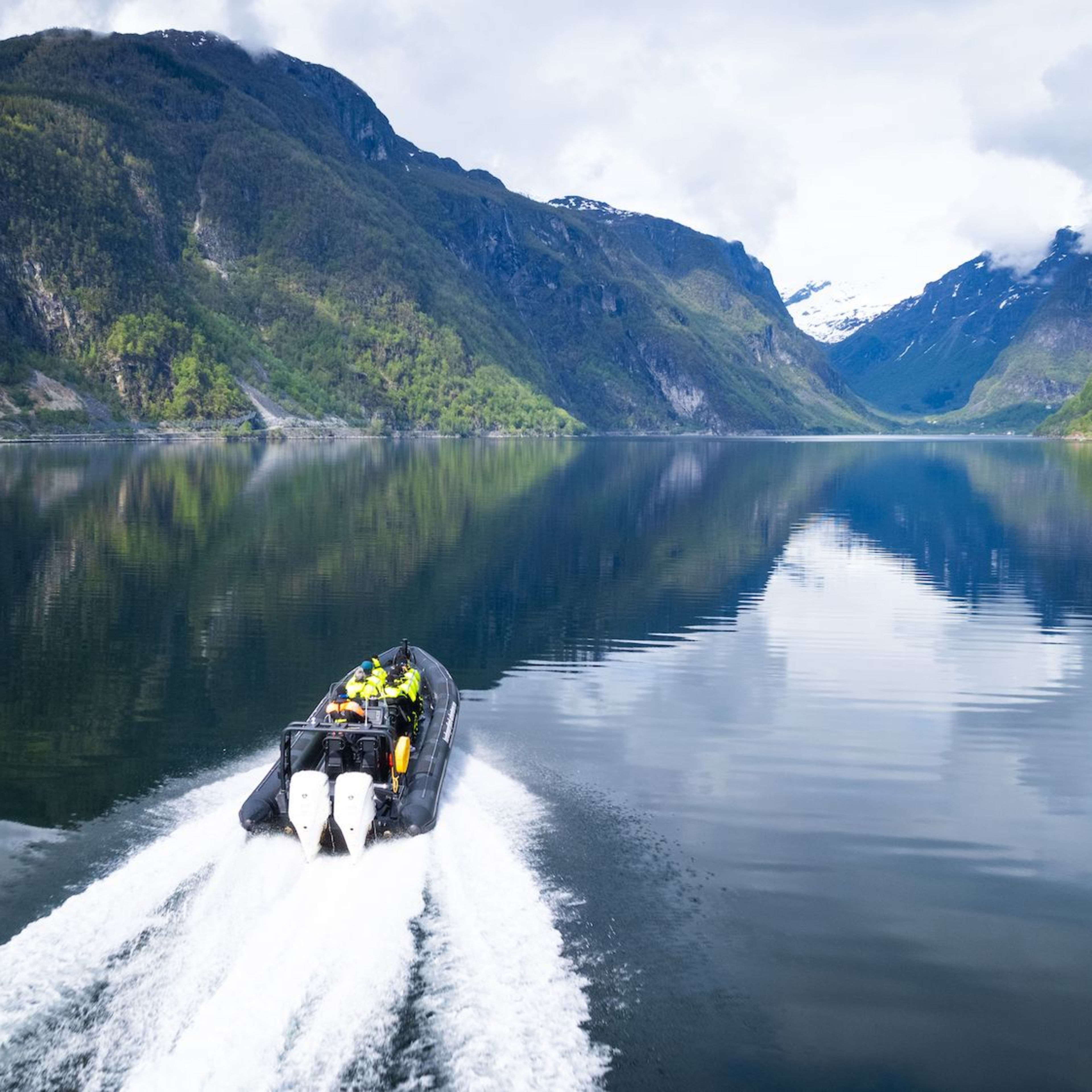 Boat with passengers in yellow jackets speeds on reflective fjord surrounded by steep forested mountains.