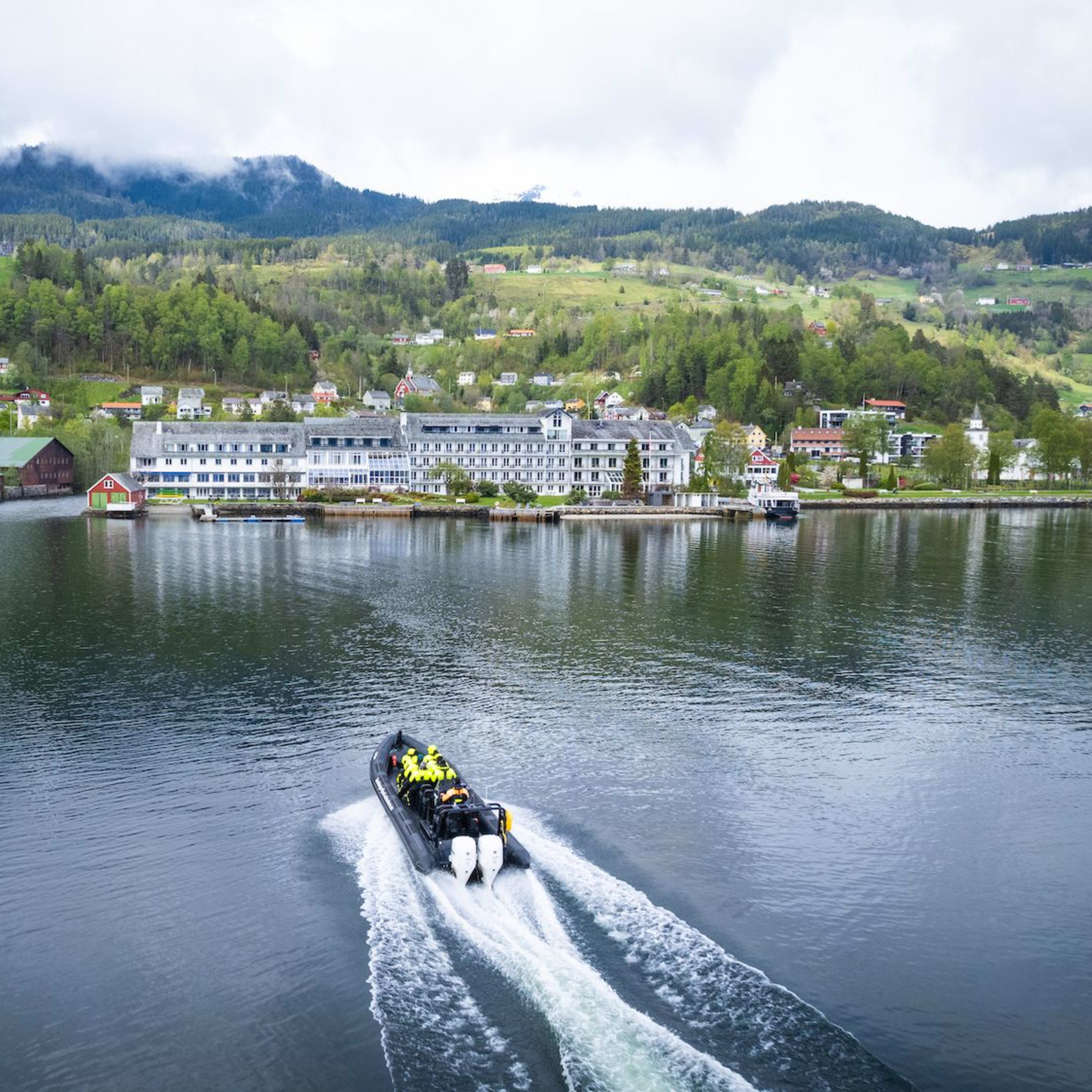 Boat with passengers in yellow jackets passes charming village with colorful houses along reflective fjord.