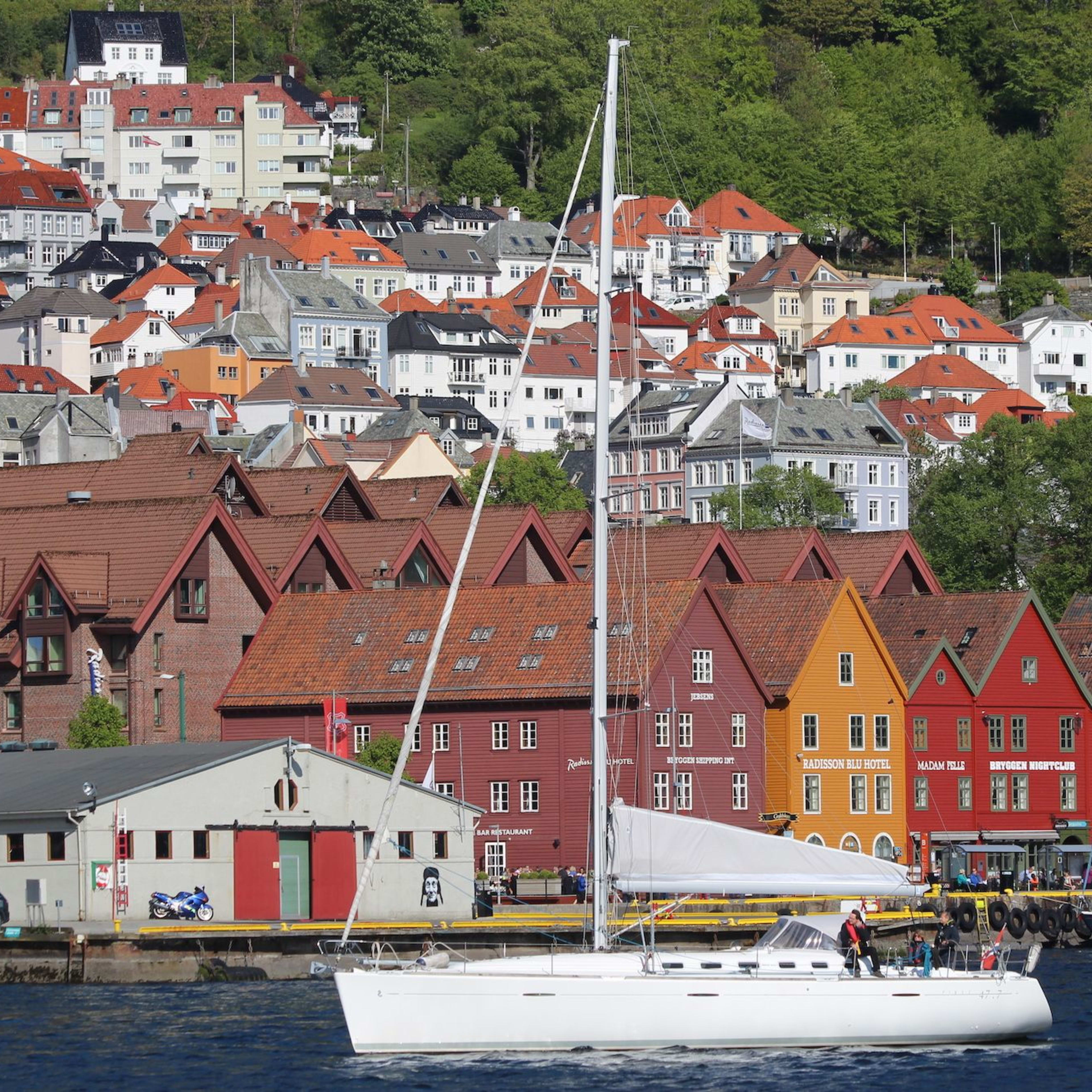 Weißes Segelboot mit gesenktem Segel liegt nahe den bunten Bryggen-Gebäuden in Bergen, mit charmanten Hütten am Hügel im Hintergrund.