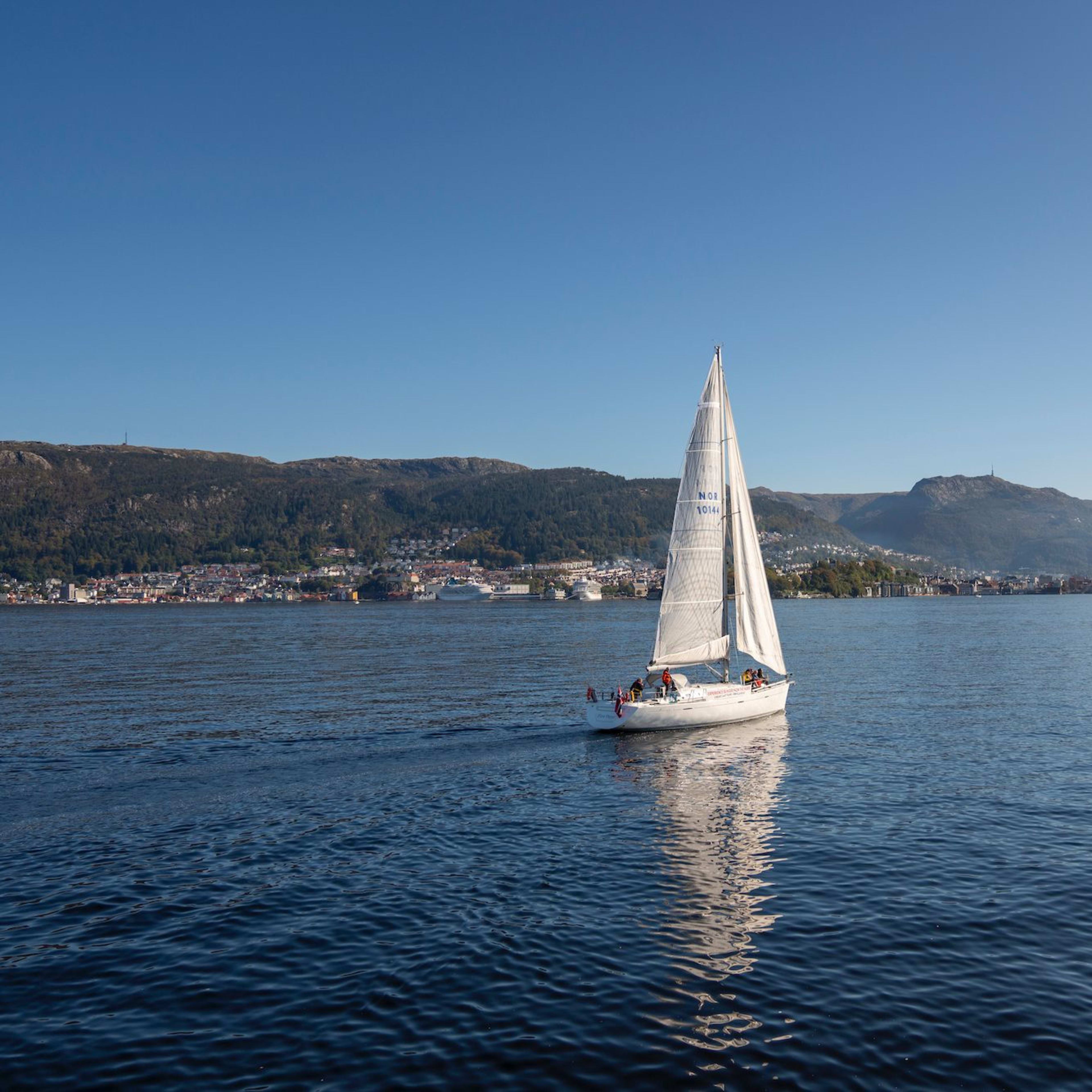 Weißes Segelboot mit "NOR 1014-4" Segel auf ruhigem, tiefblauem Wasser unter klarem Himmel, mit Küstenstadt im Hintergrund.