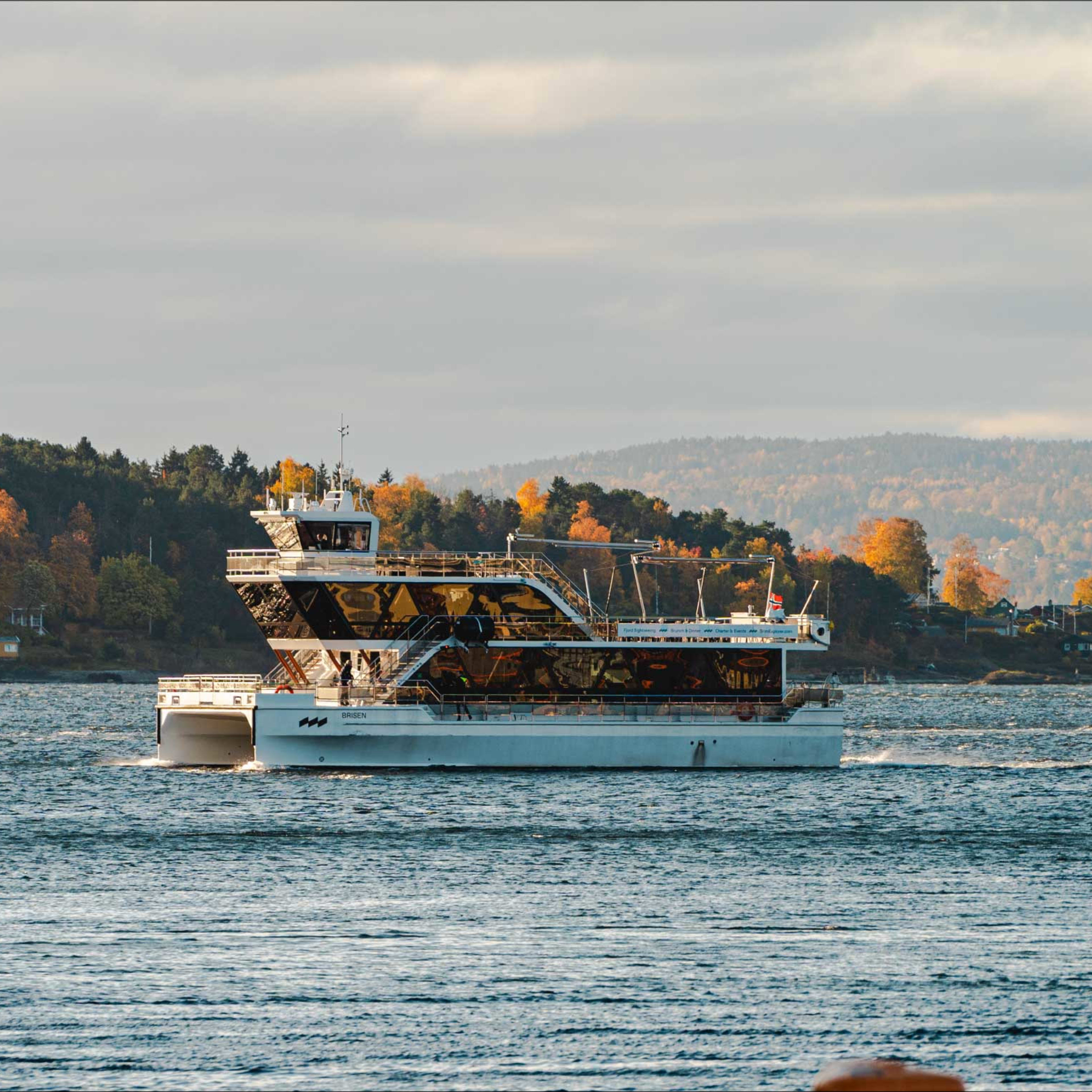 Hvit hybrid-elektrisk cruisebåt seiler gjennom Oslofjordens farvann med høstfargede skogåser