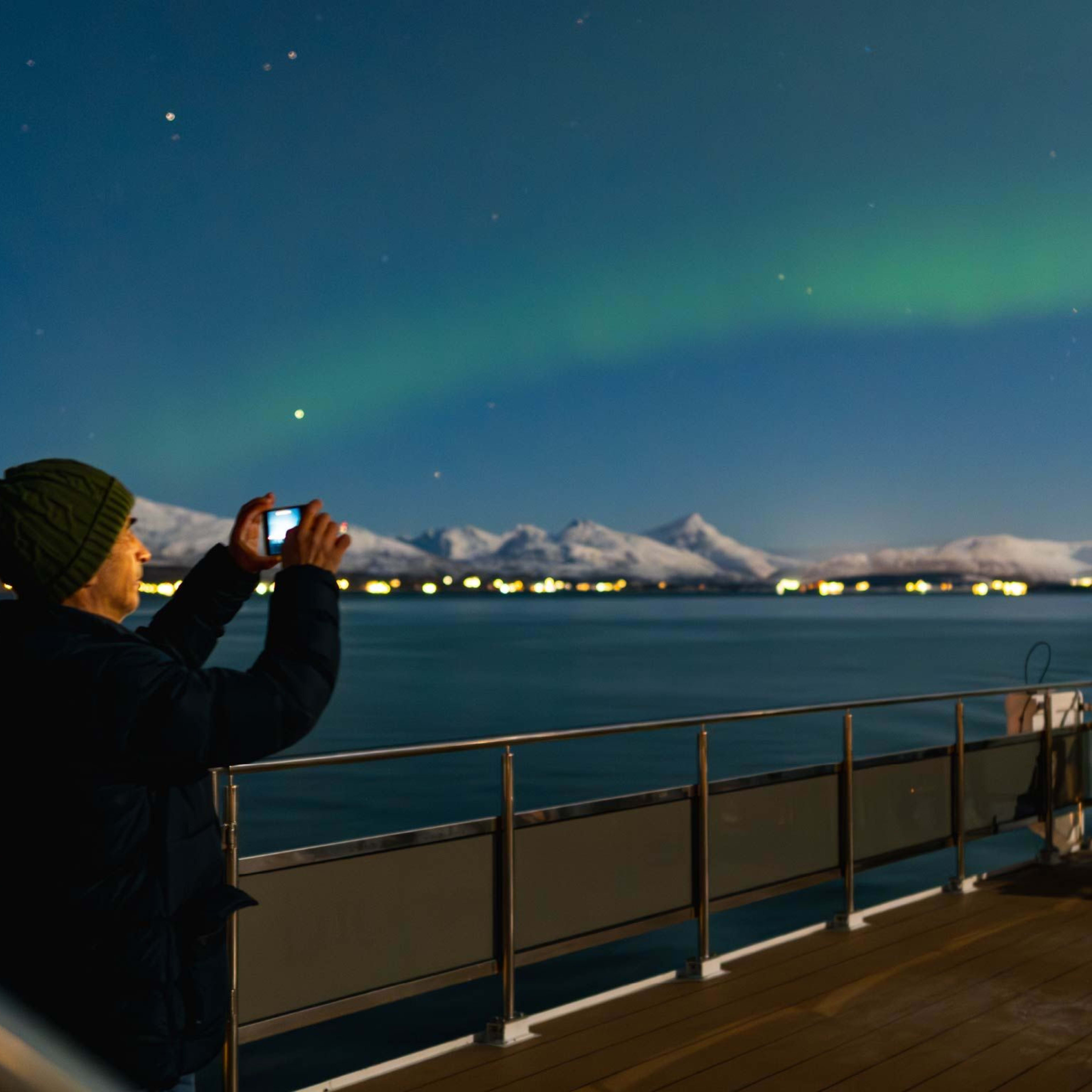 En person på skipets dekk fanger nordlyset over snødekte fjell, med grønne auroraer som lyser opp nattehimmelen.