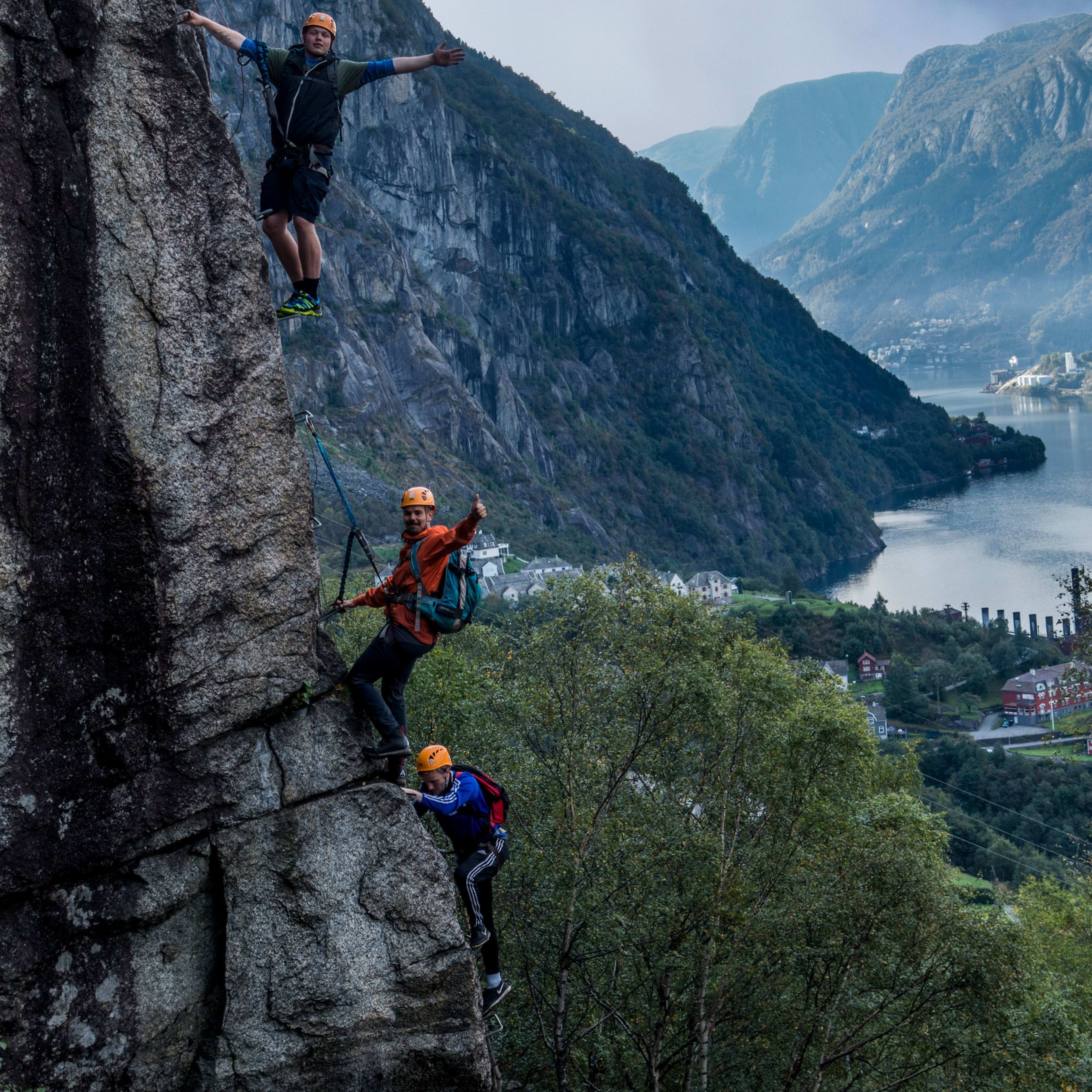 Kletterer hebt Arme an Felswand mit nebligem Fjord.
