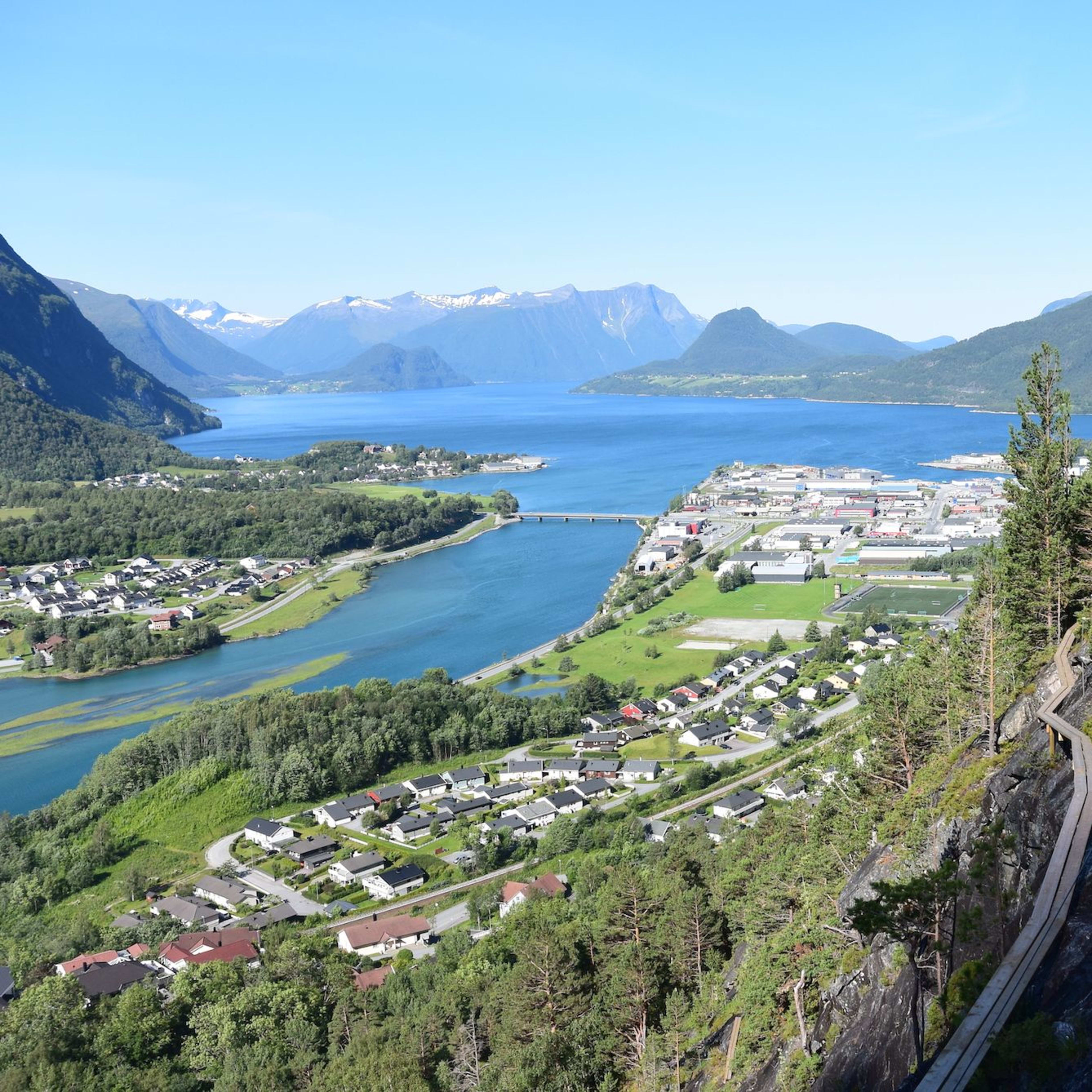 Luftaufnahme einer Küstenstadt zwischen Bergen und Fjord, mit Holzweg, Brücke und grünen Feldern.