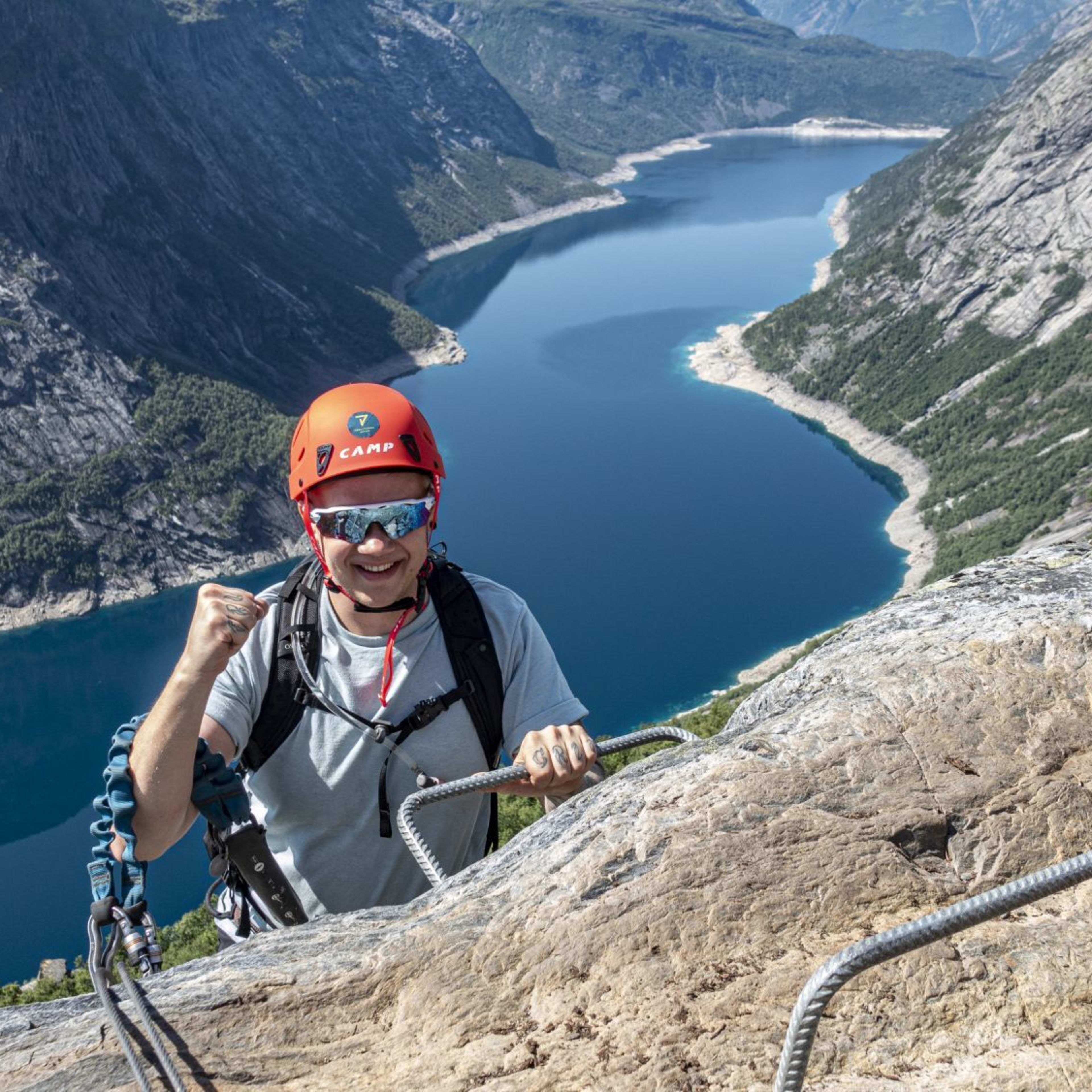 Climber at top of rock face celebrating with raised fist, wearing orange helmet and sunglasses, lake and mountains in background.