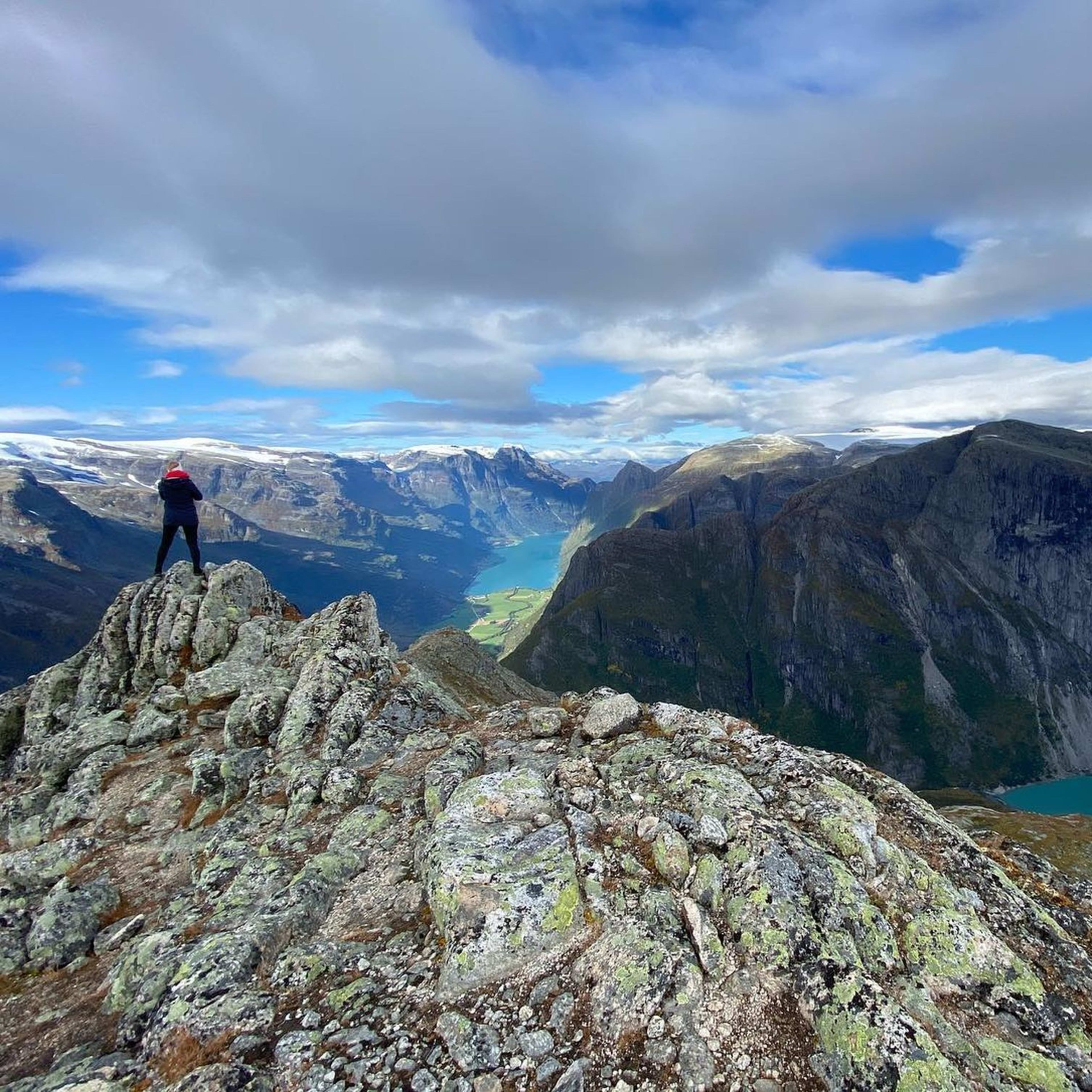 Person, die triumphierend auf einem Felsen steht, mit Blick auf den Fjord und schneebedeckte Berge im Hintergrund.