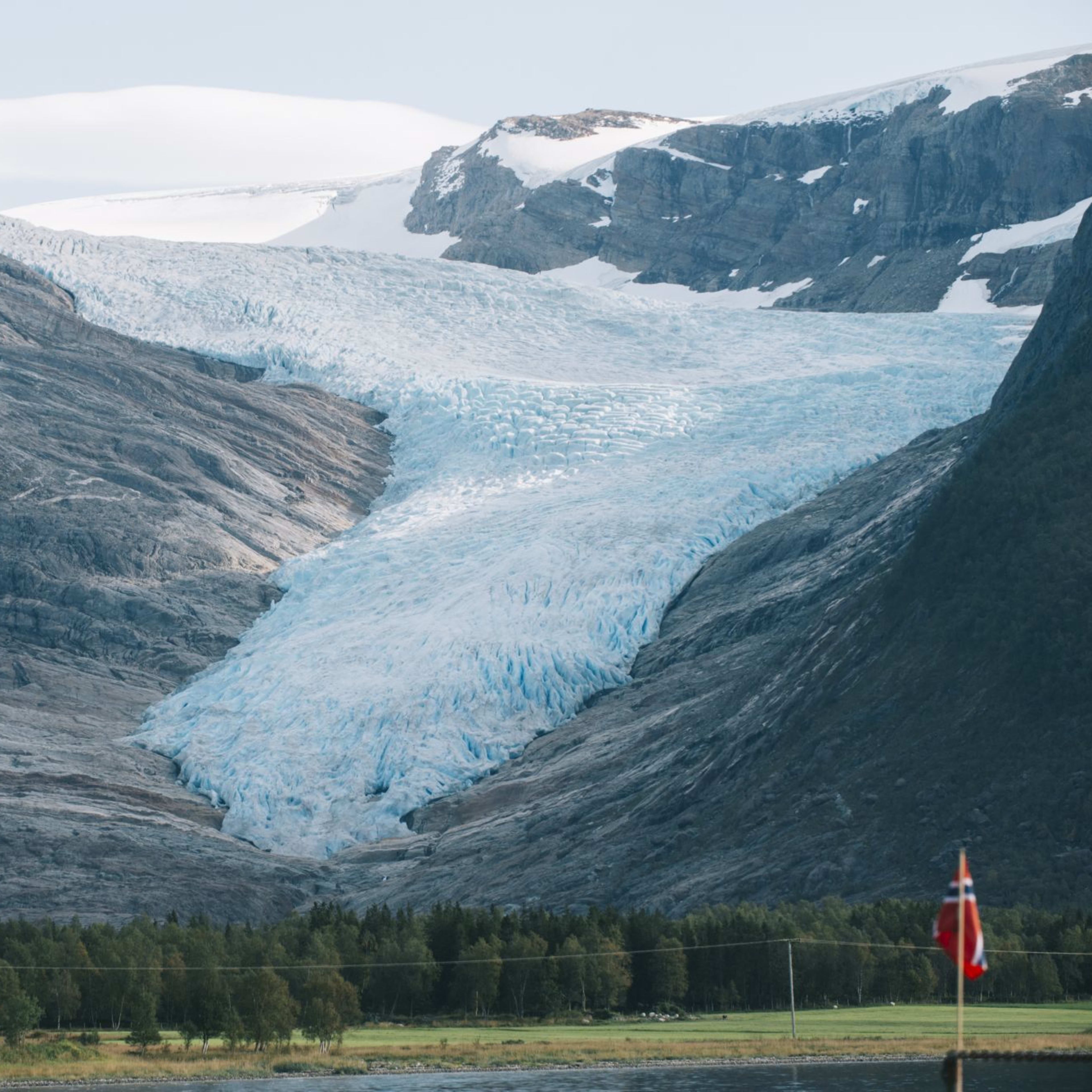 Atemberaubende Aussicht auf den Gletscher, der von den Bergen hinabfließt, mit einer Flagge im Vordergrund, grünen Bäumen und schneebedeckten Gipfeln.