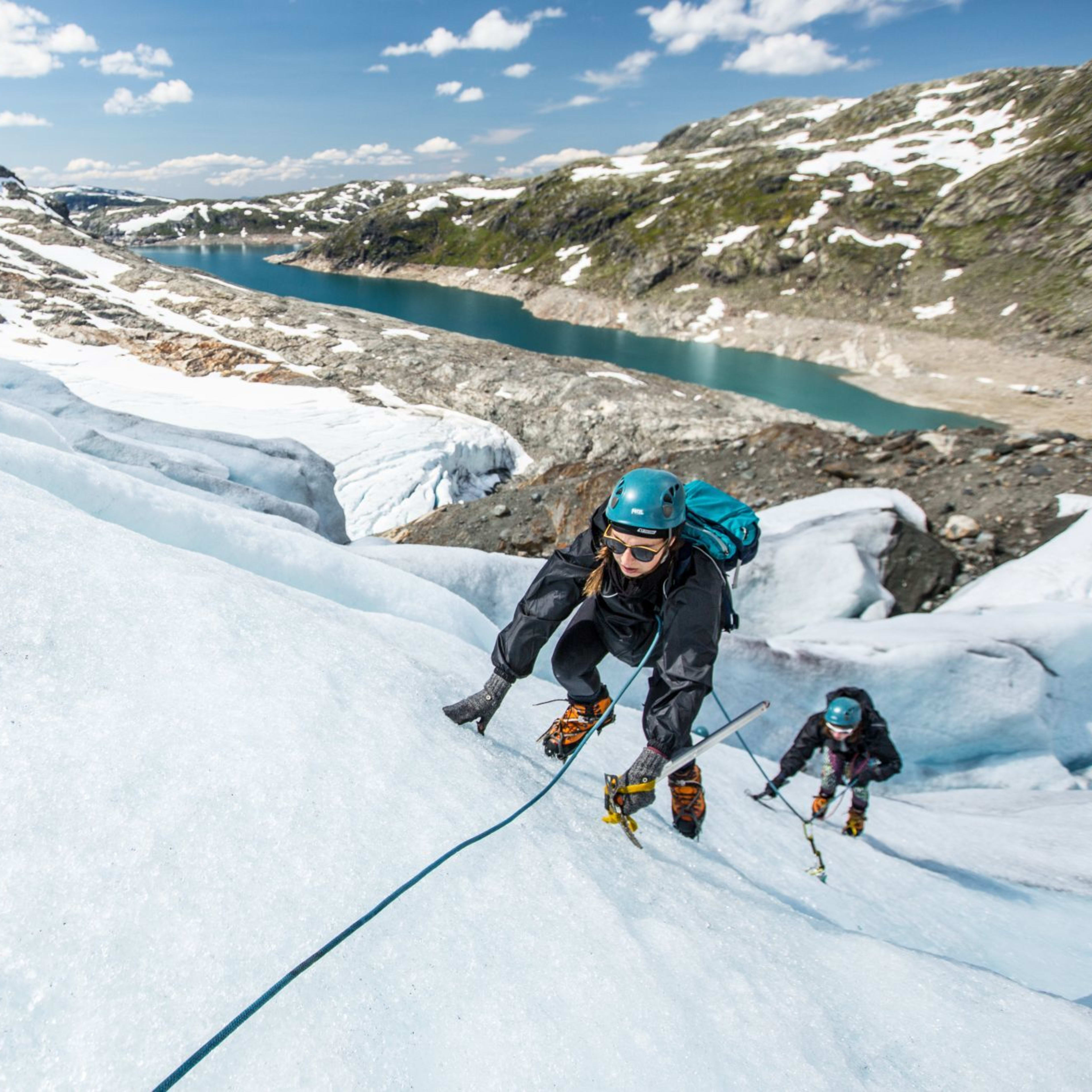 En klatrer bestiger en isbre med en isøks og tau, mens andre er synlige i bakgrunnen nær en fjord.