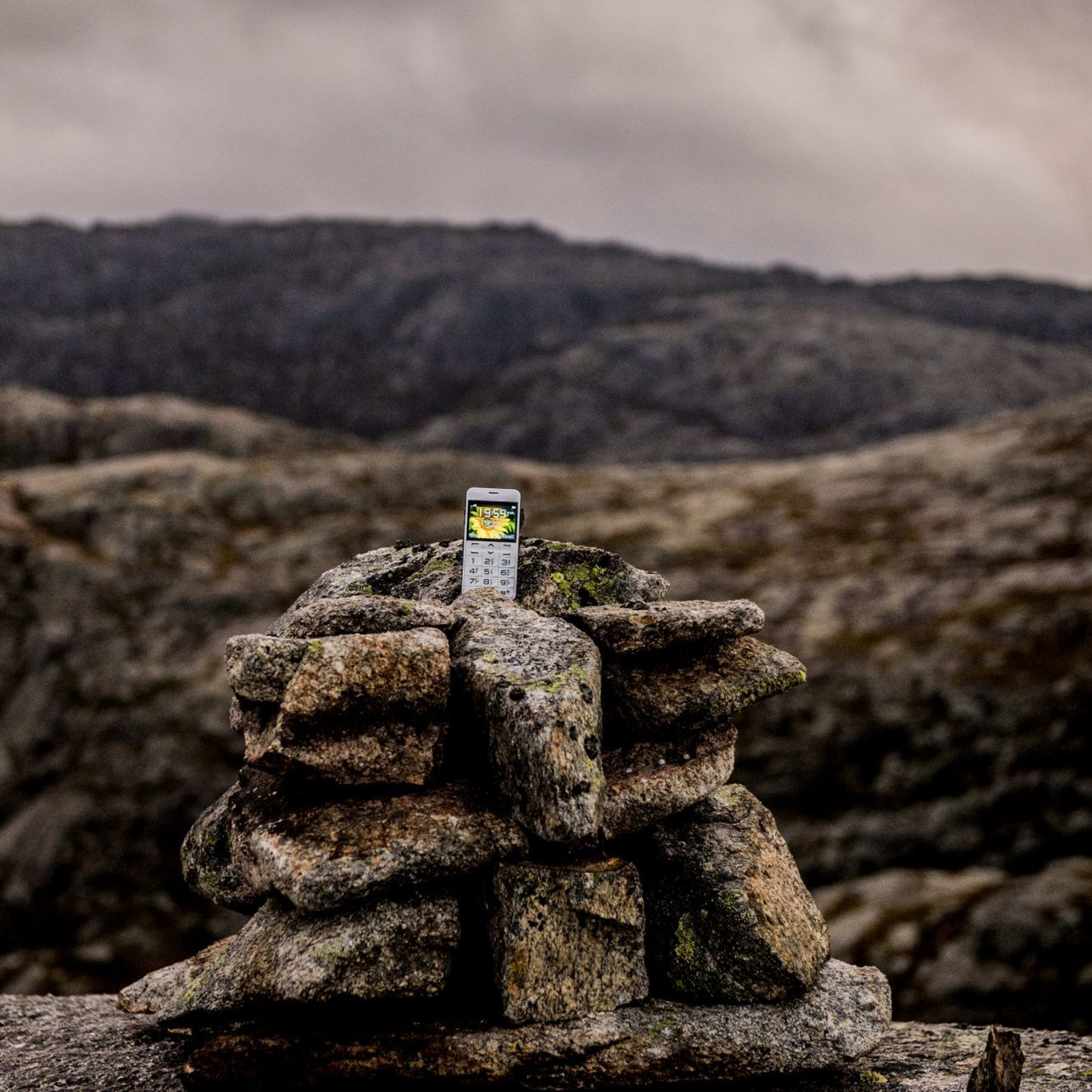 Old mobile phone placed on stone cairn against rugged mountain landscape and cloudy sky.
