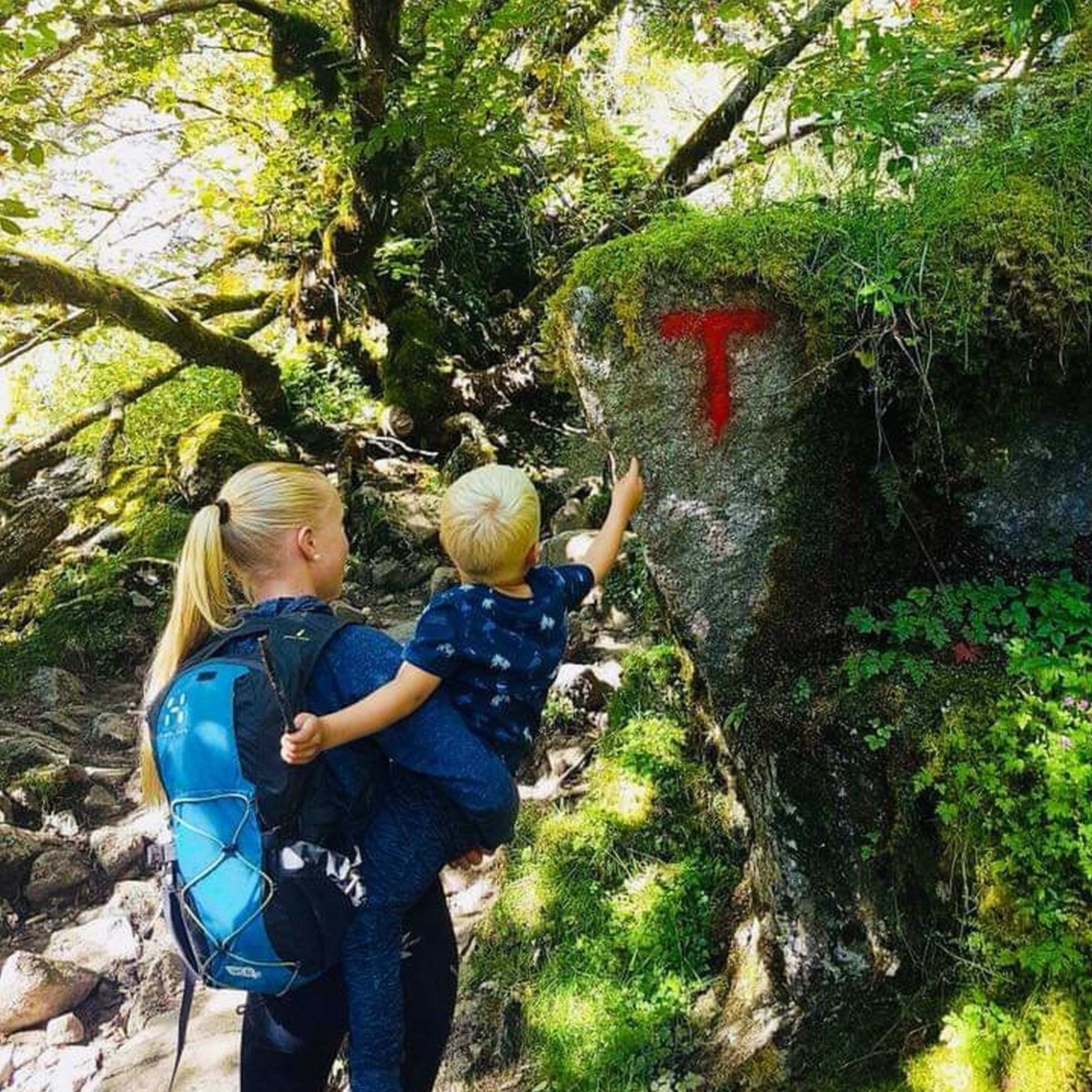 Kvinne som bærer barn, går gjennom skogen og peker på et stisyne med rød "T"-symbol.