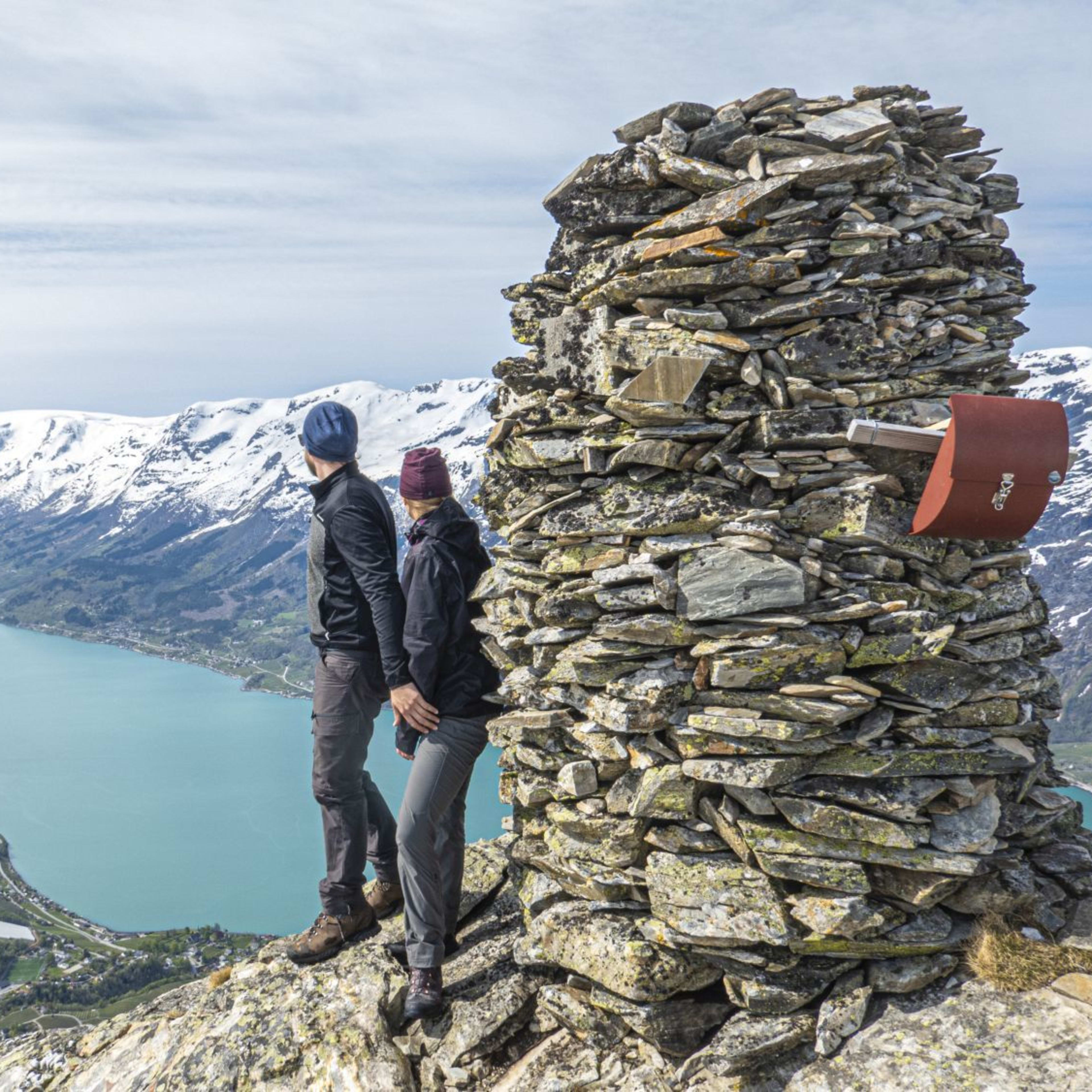 Two hikers standing by tall cairn overlooking turquoise fjord, with snow-capped mountains and bright sky.