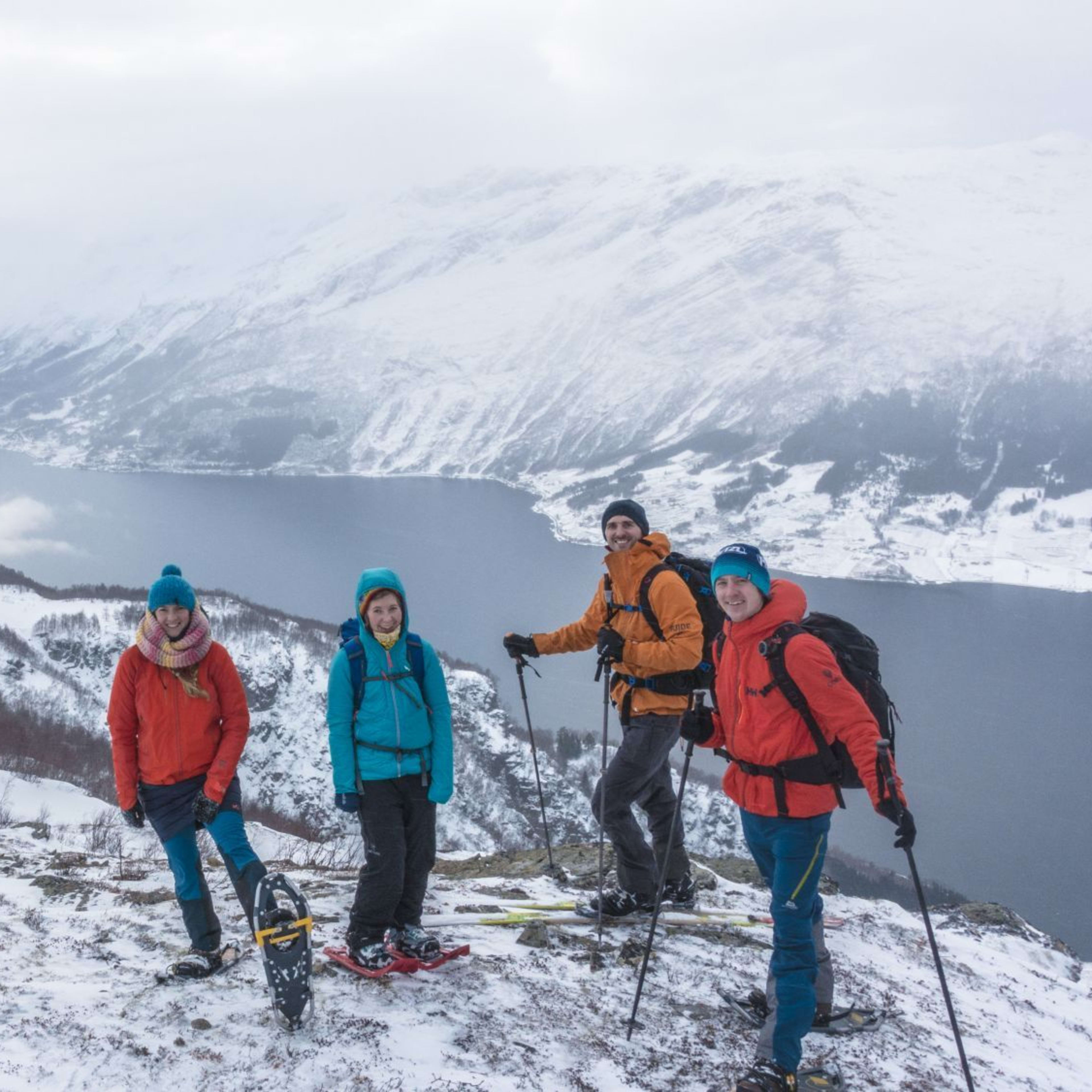Four hikers smiling on snowy slope overlooking serene fjord with snow-covered mountains under cloudy sky.