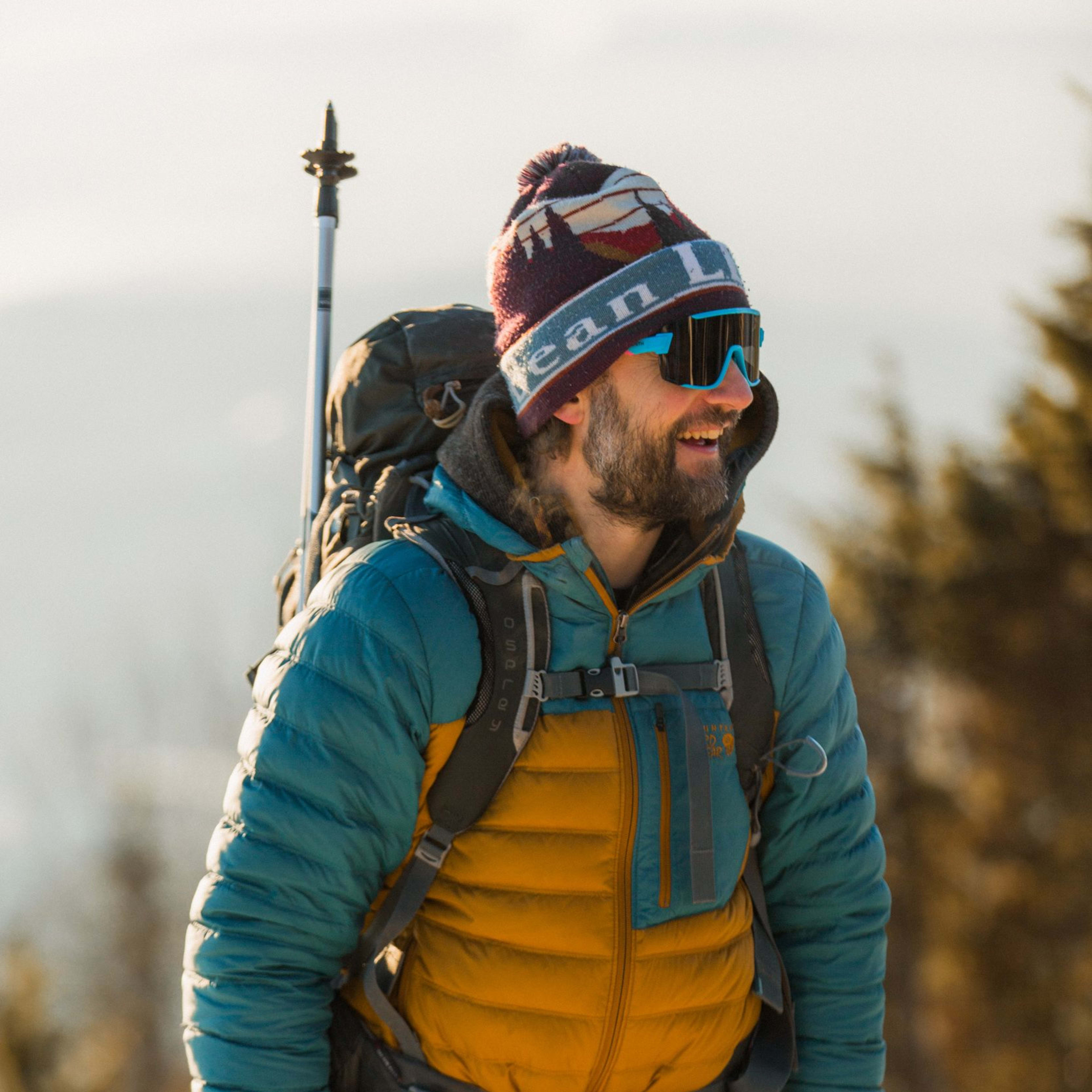 Smiling hiker in colorful winter gear with backpack and sunglasses on snowy mountain trail.