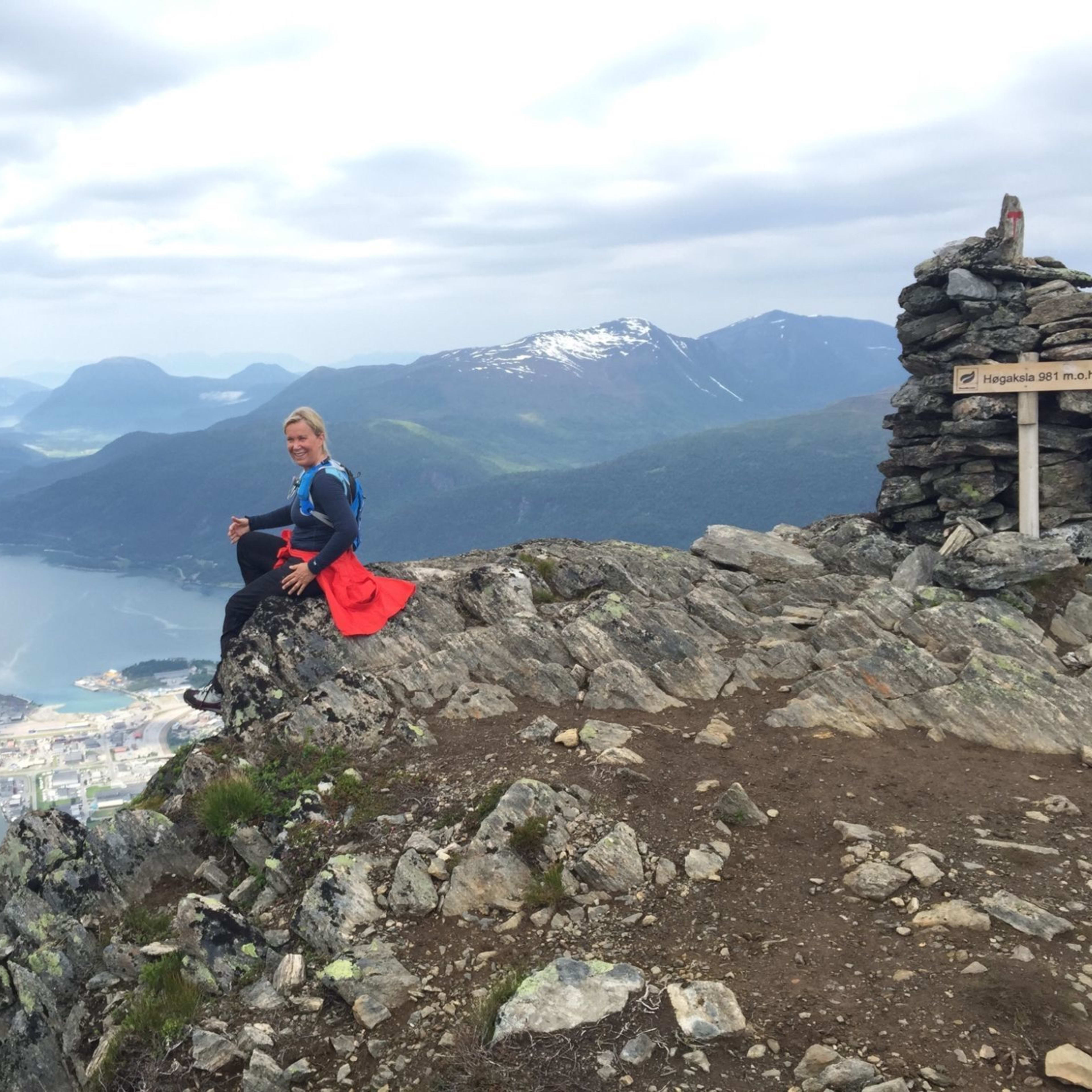 Lachende Frau sitzt an einer Klippe neben dem Schild „Høgaksla 981 m.o.h.“, mit Stadt darunter.