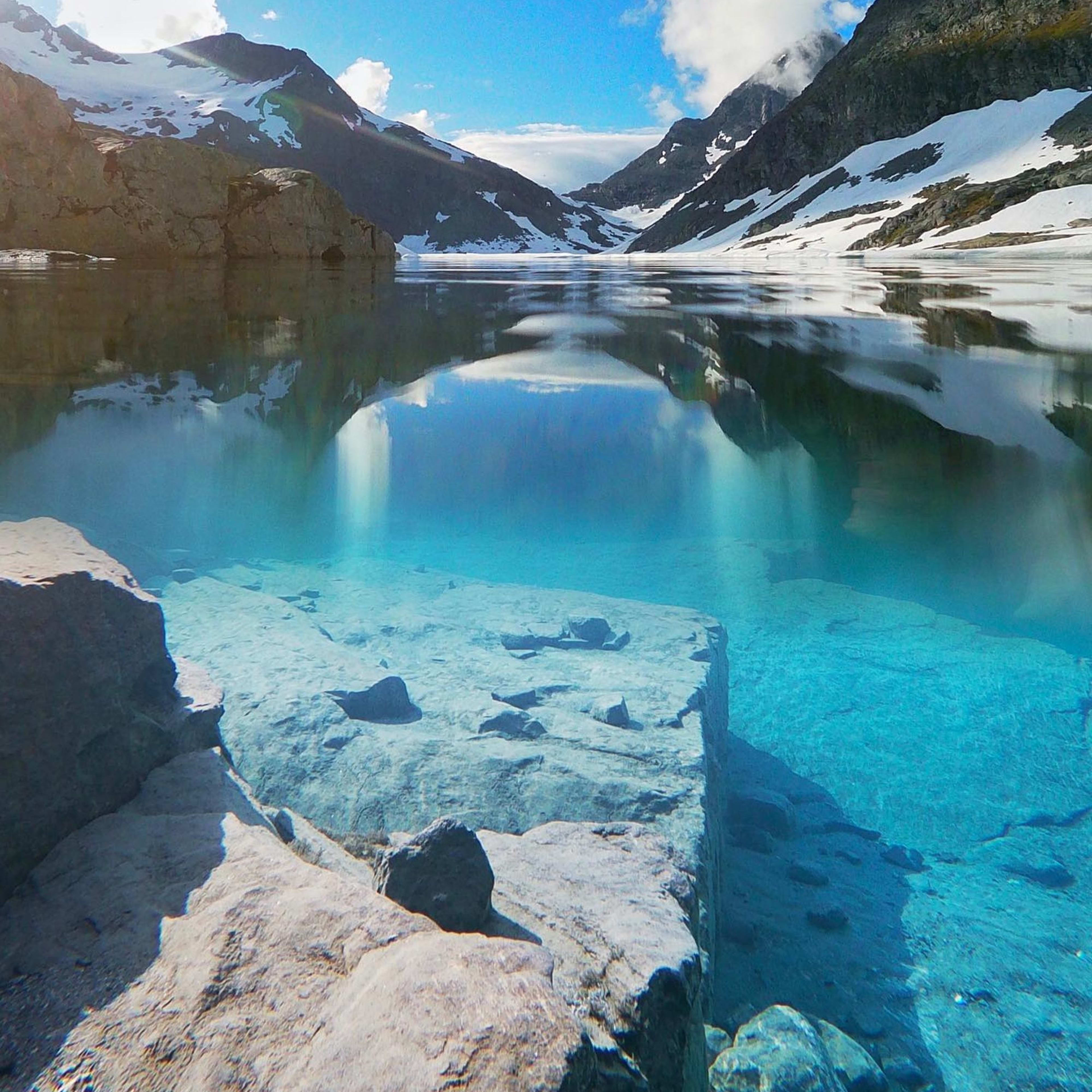 Ruhiger Bergsee mit klarem Wasser, das die umliegenden Gipfel und den blauen Himmel widerspiegelt, mit Felsen am Ufer.