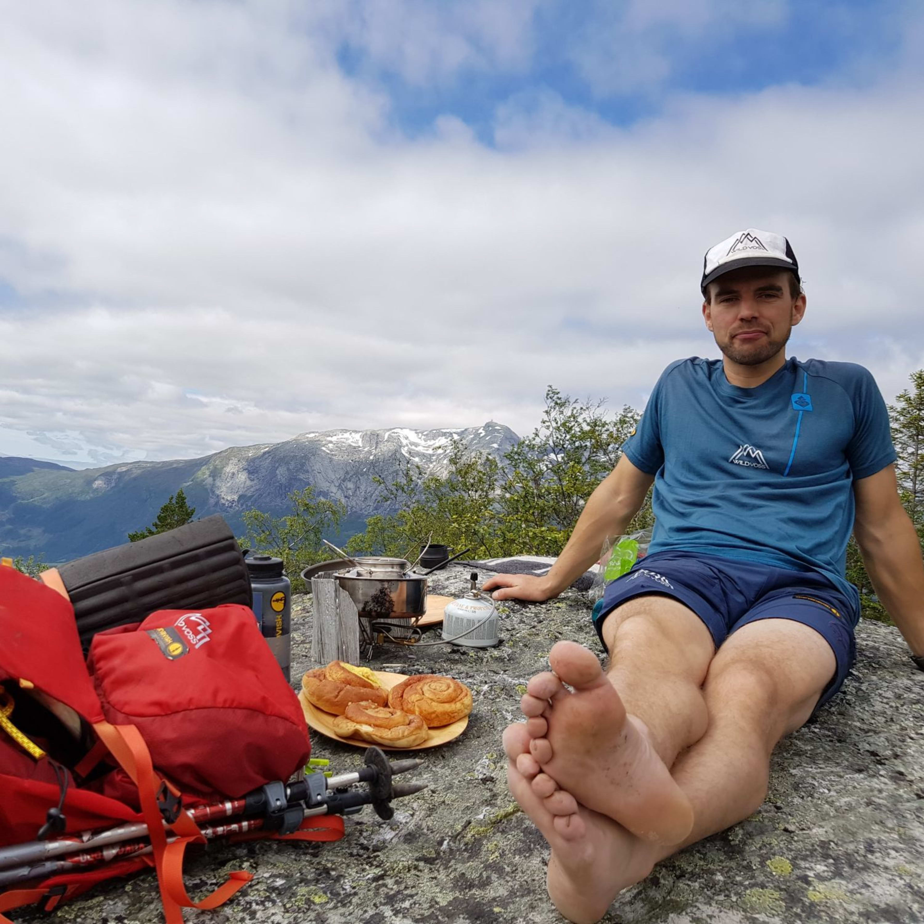 Mann som hviler på en steinete overflate under tur, og nyter en piknik med bakverk og en liten campingovn, med vakre fjell i bakgrunnen.