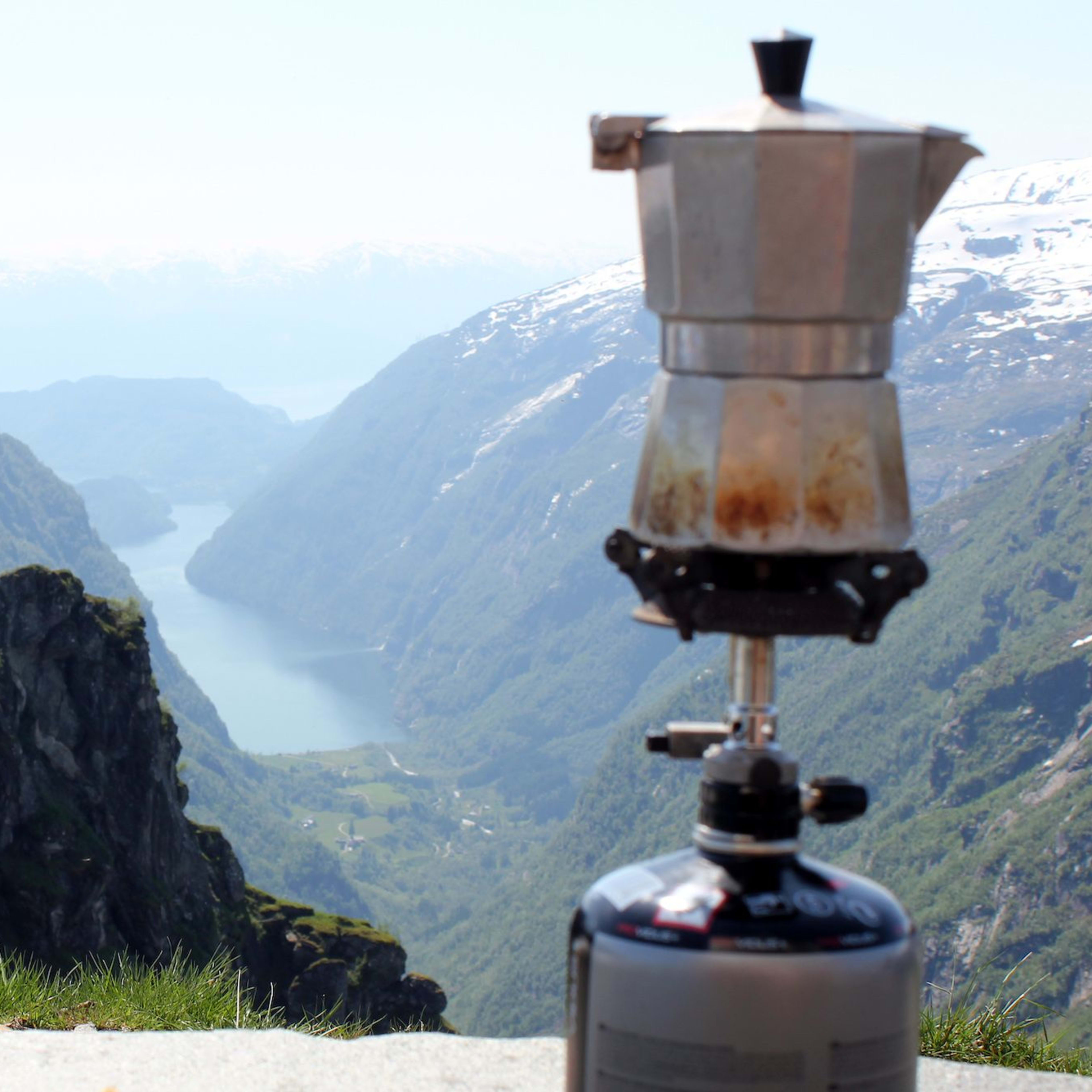 Metallmoka på portable gasskomfyr brygger kaffe med fjord og robuste fjell i bakgrunnen.