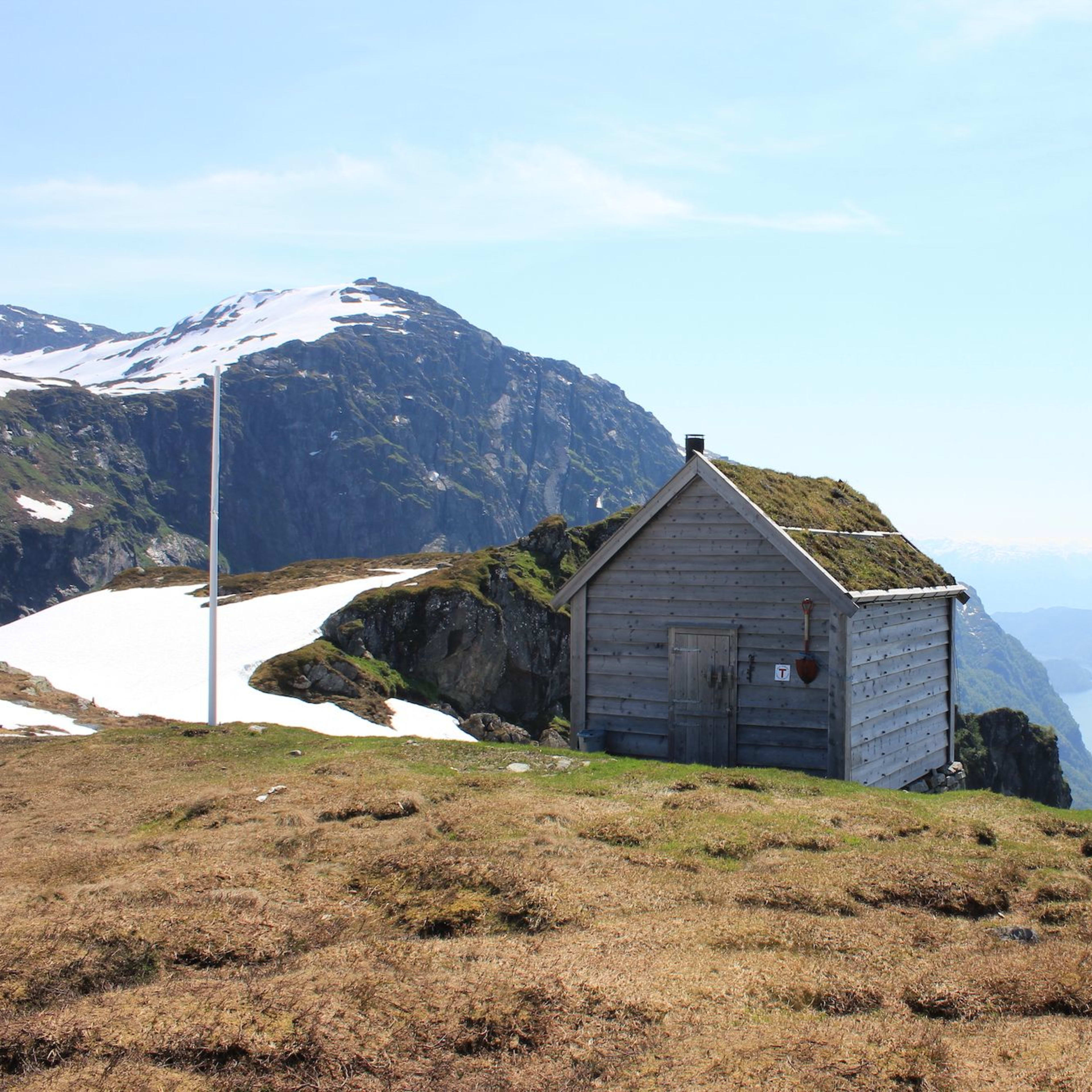 Liten trehytte på en gresskledd høyde med snøflekker, med utsikt over en svingende fjord og bratte fjell.