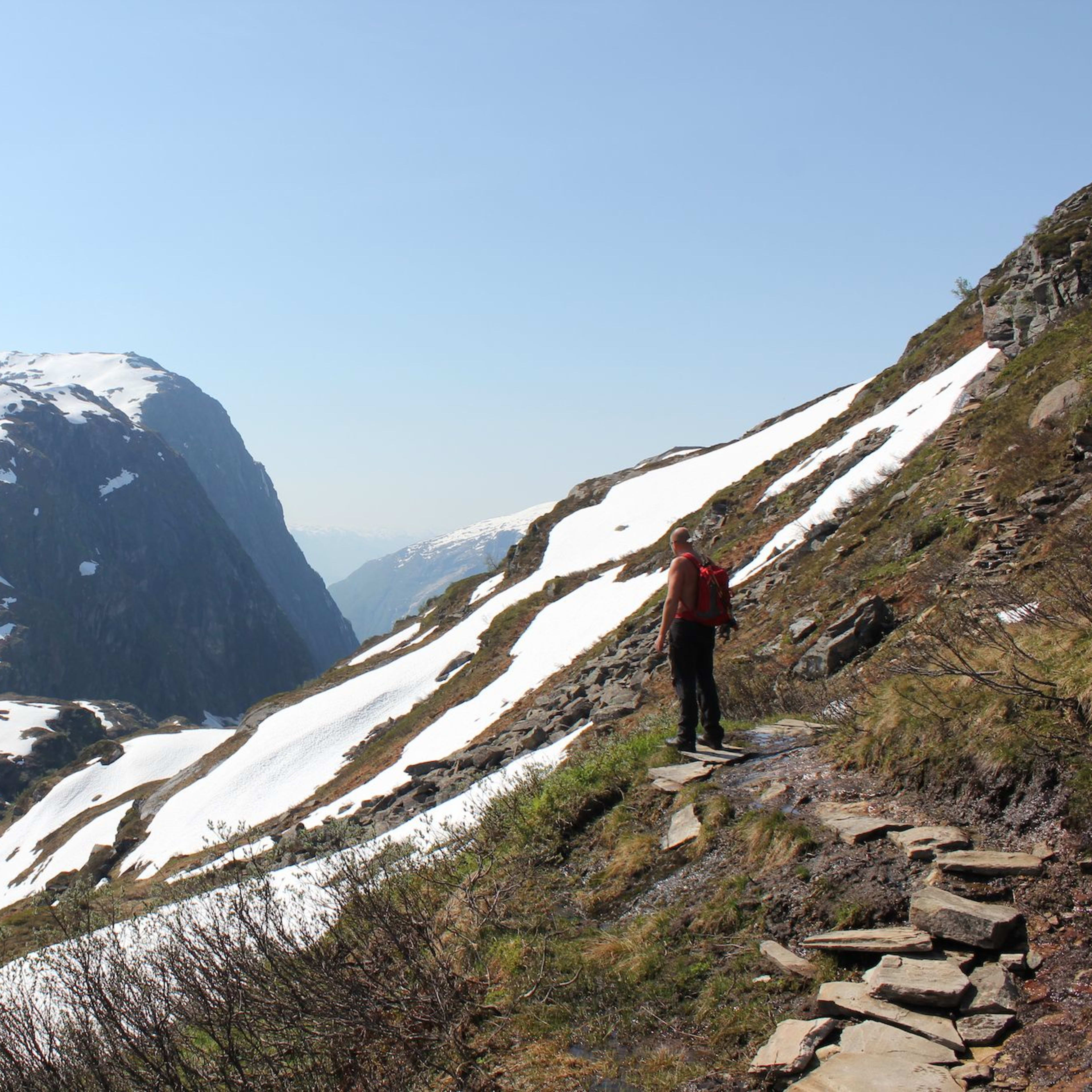 Turgåer med rød ryggsekk på en steinete fjellsti, med utsikt over dype daler og snøkledde topper.