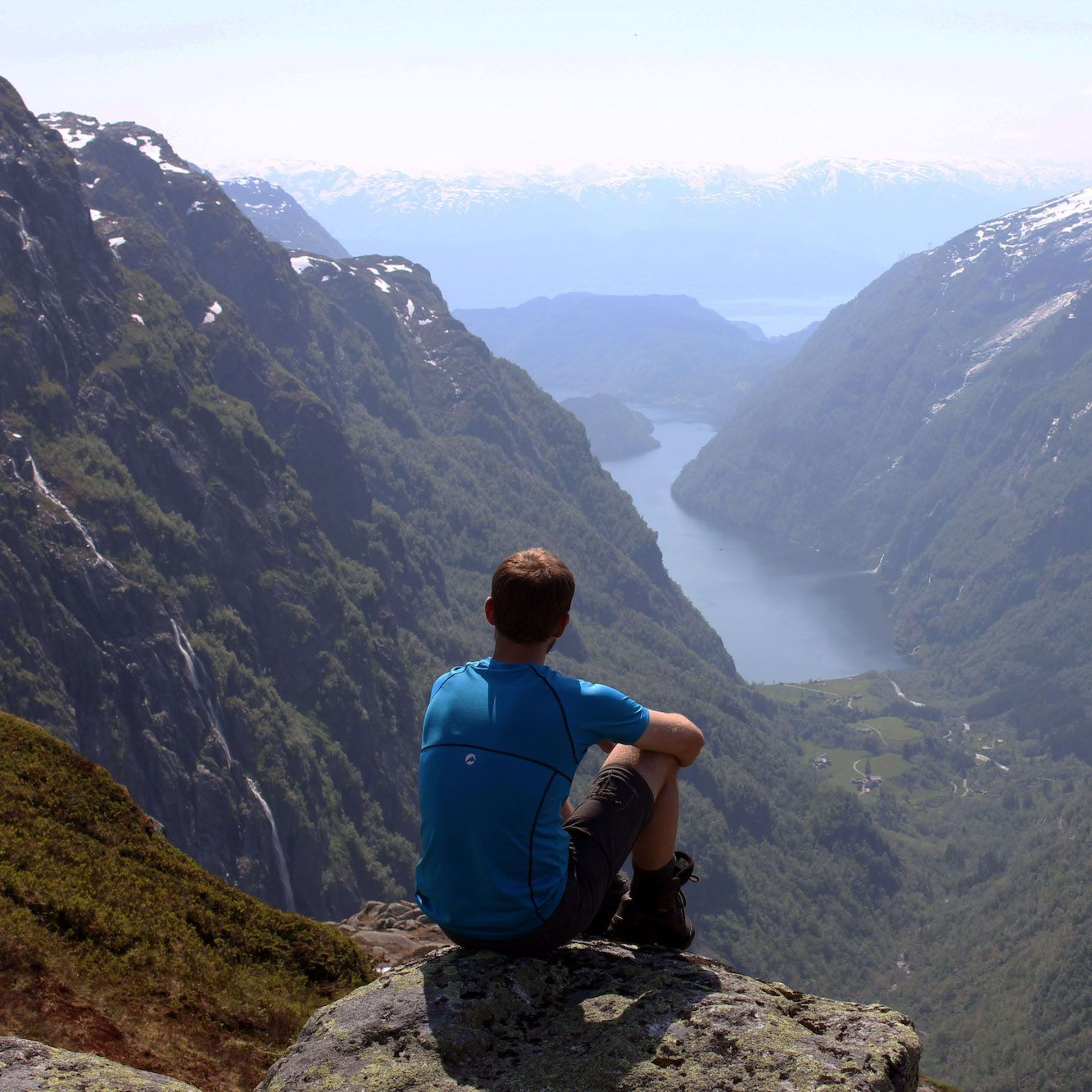 Mann som sitter på en stein og ser på en fantastisk dal med en svingende blå fjord og tårnende fjell.