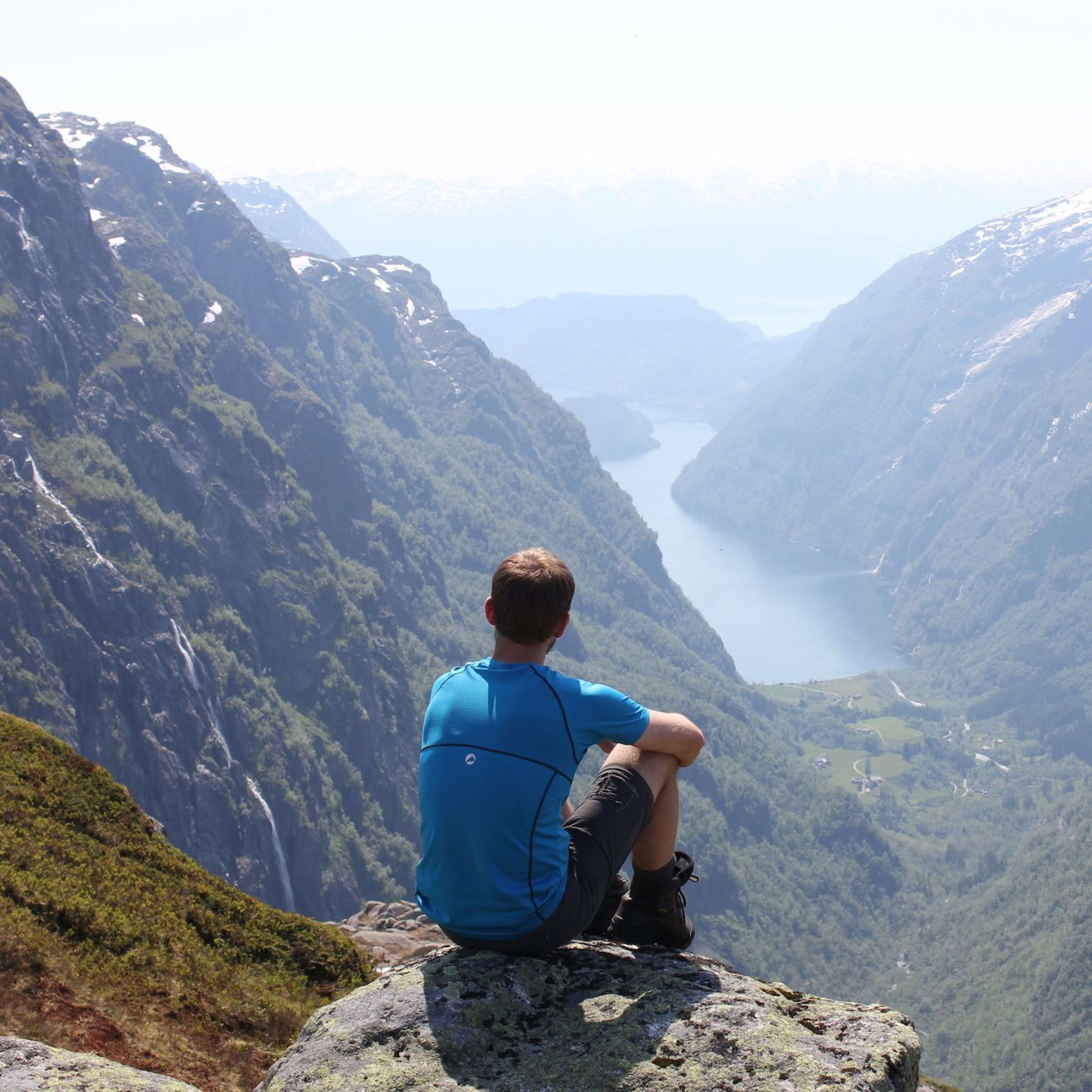 Mann som sitter på en steinete utstikker og ser på en fantastisk dal med en svingende fjord og bratte, skogkledde fjell.