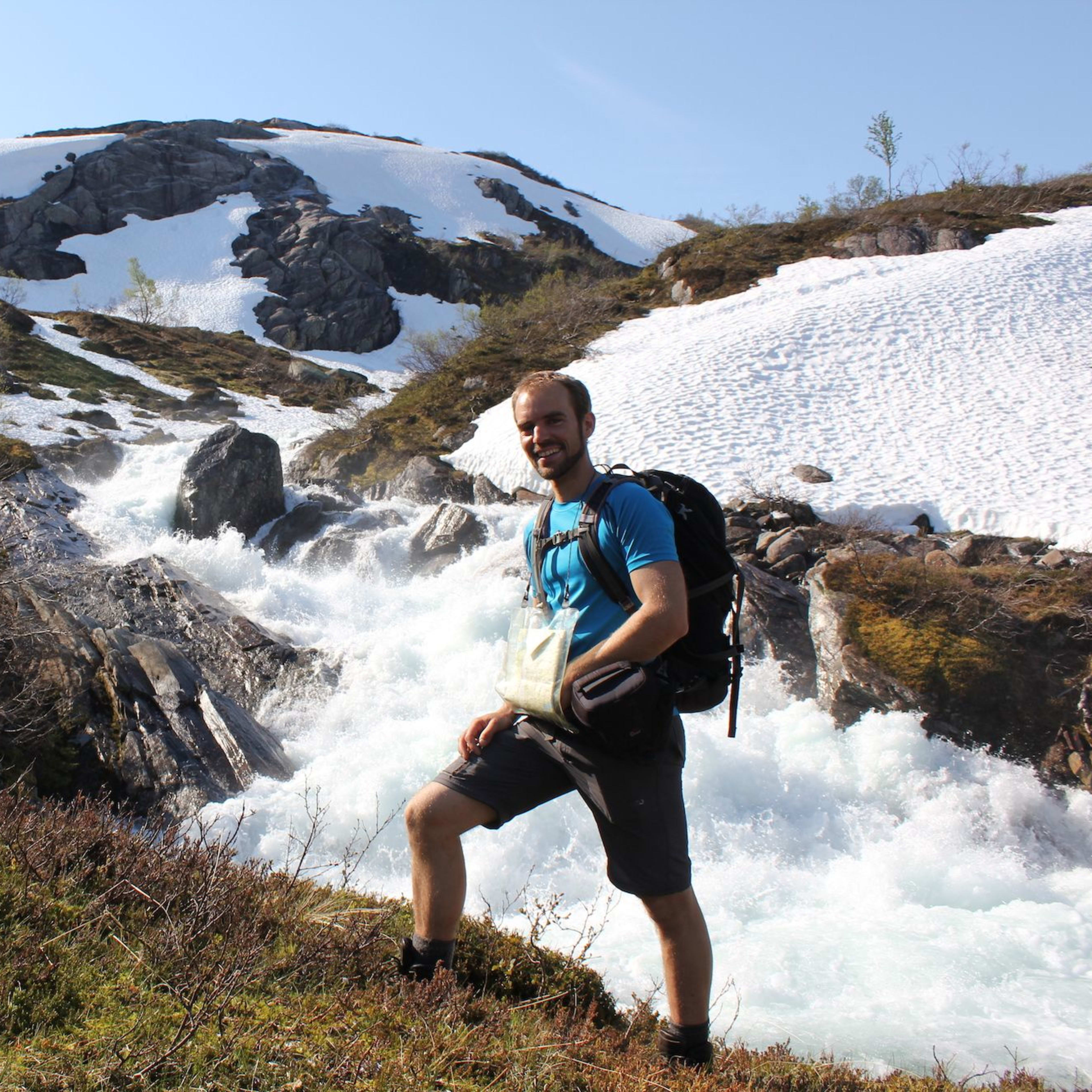 Smilende turgåer i blå skjorte og ryggsekk står ved skummende fjellbekk, med røft landskap og smeltende snø.