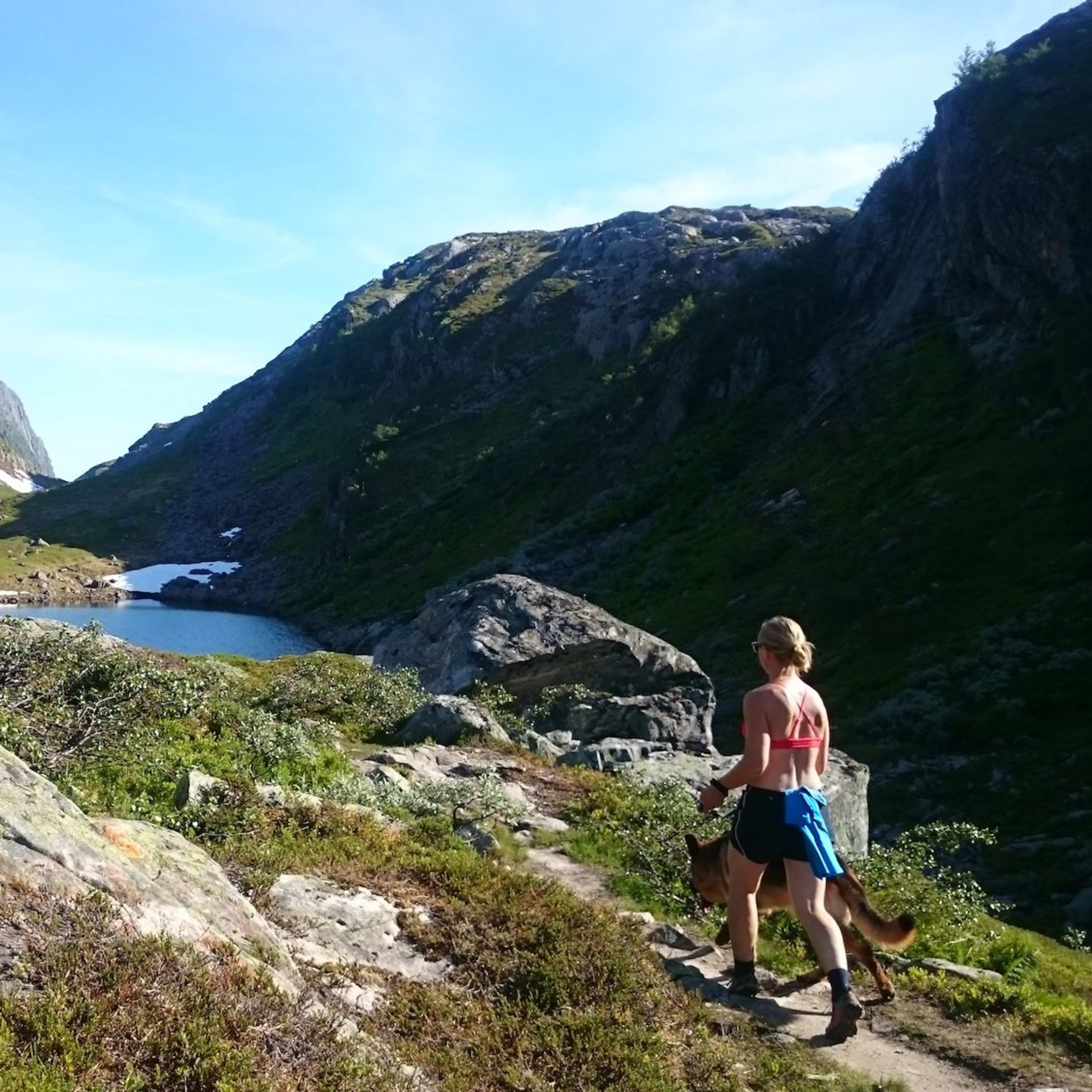 Kvinne i treningsklær går langs en steinete fjellsti med hund, nær en rolig innsjø omgitt av åser.