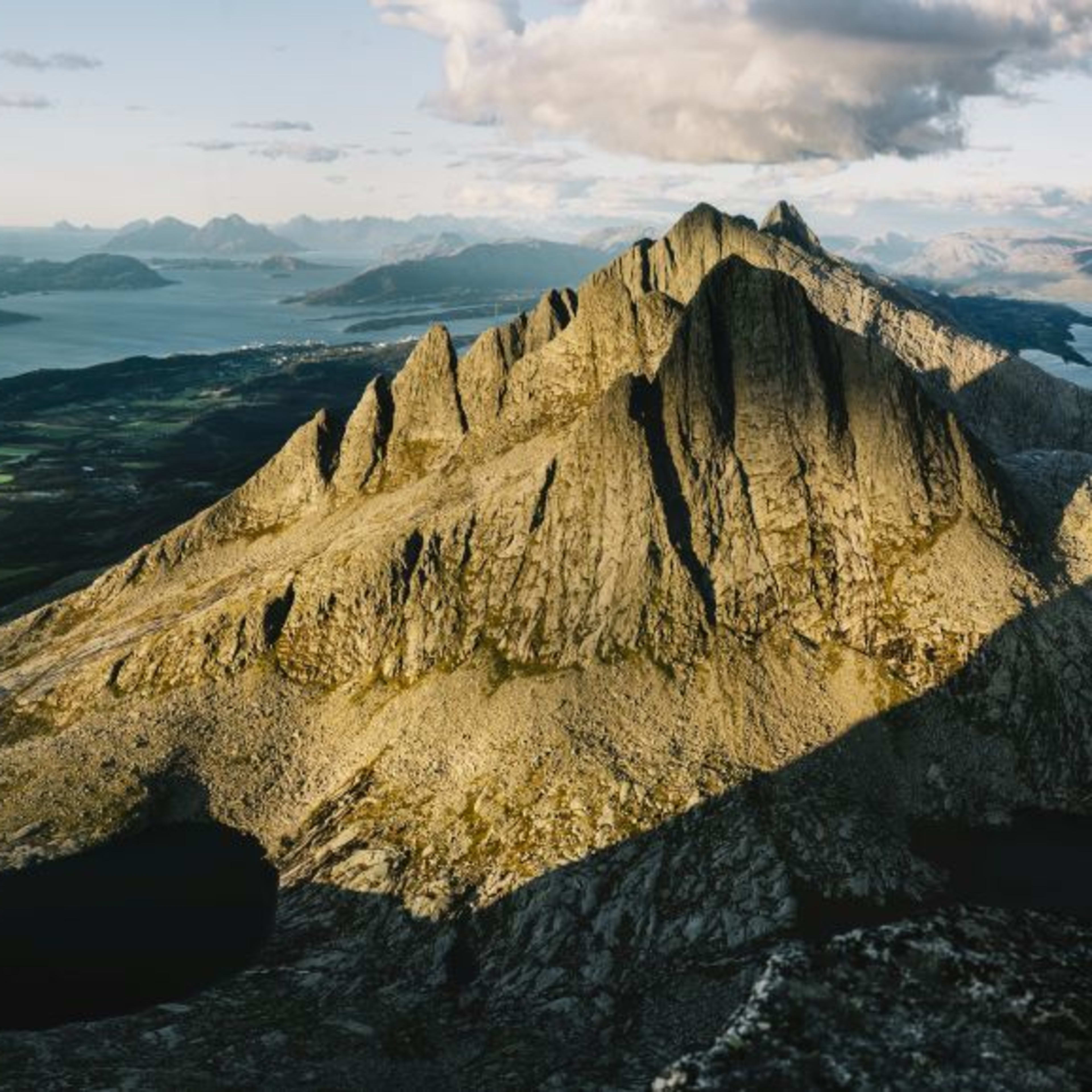 Fotograf i gul jakke som fanger kystlandskapet med dramatiske fjell, grønne jordbruksområder og rolig hav.