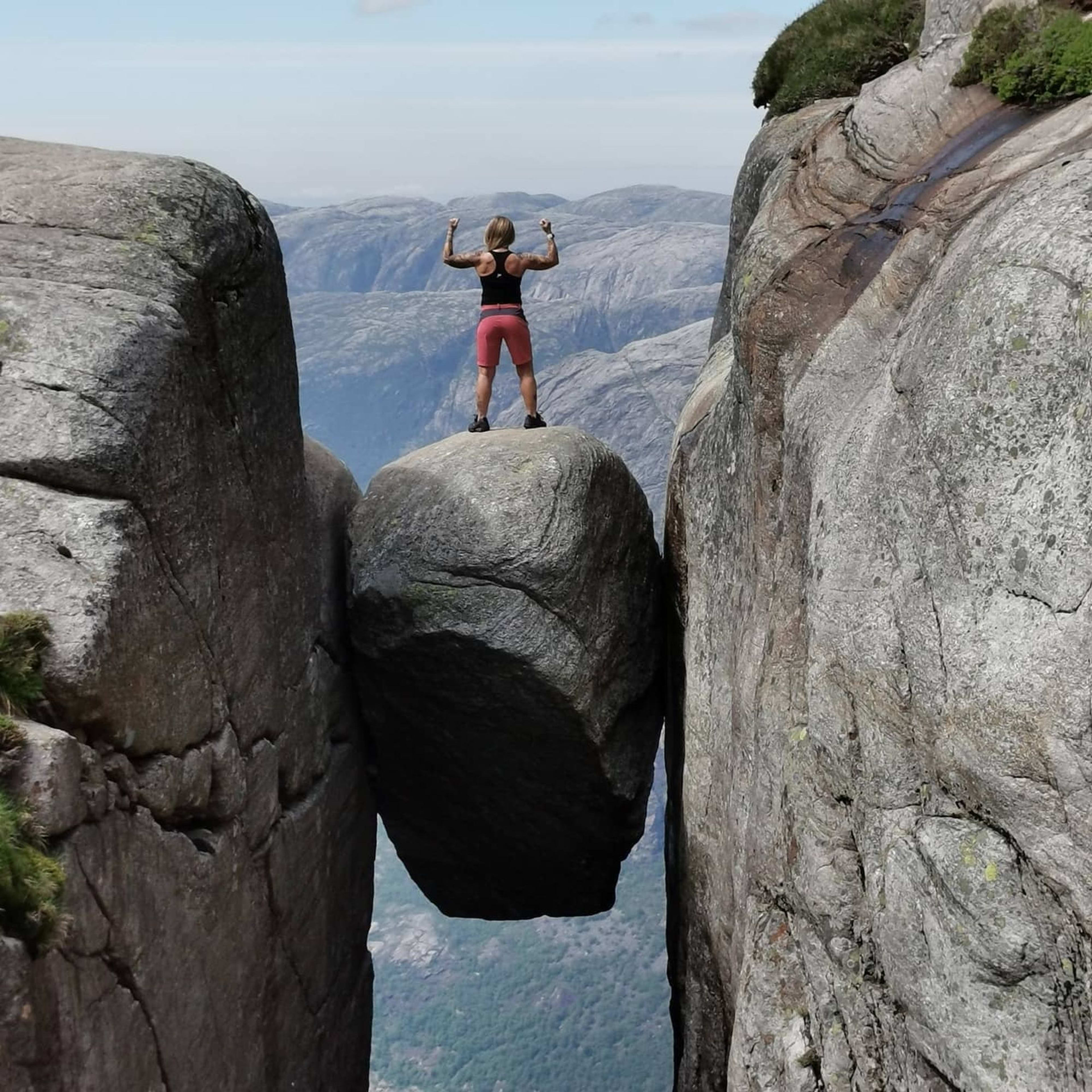 Woman standing triumphantly on Kjeragbolten, large boulder wedged between steep rock faces above deep mountain valley.