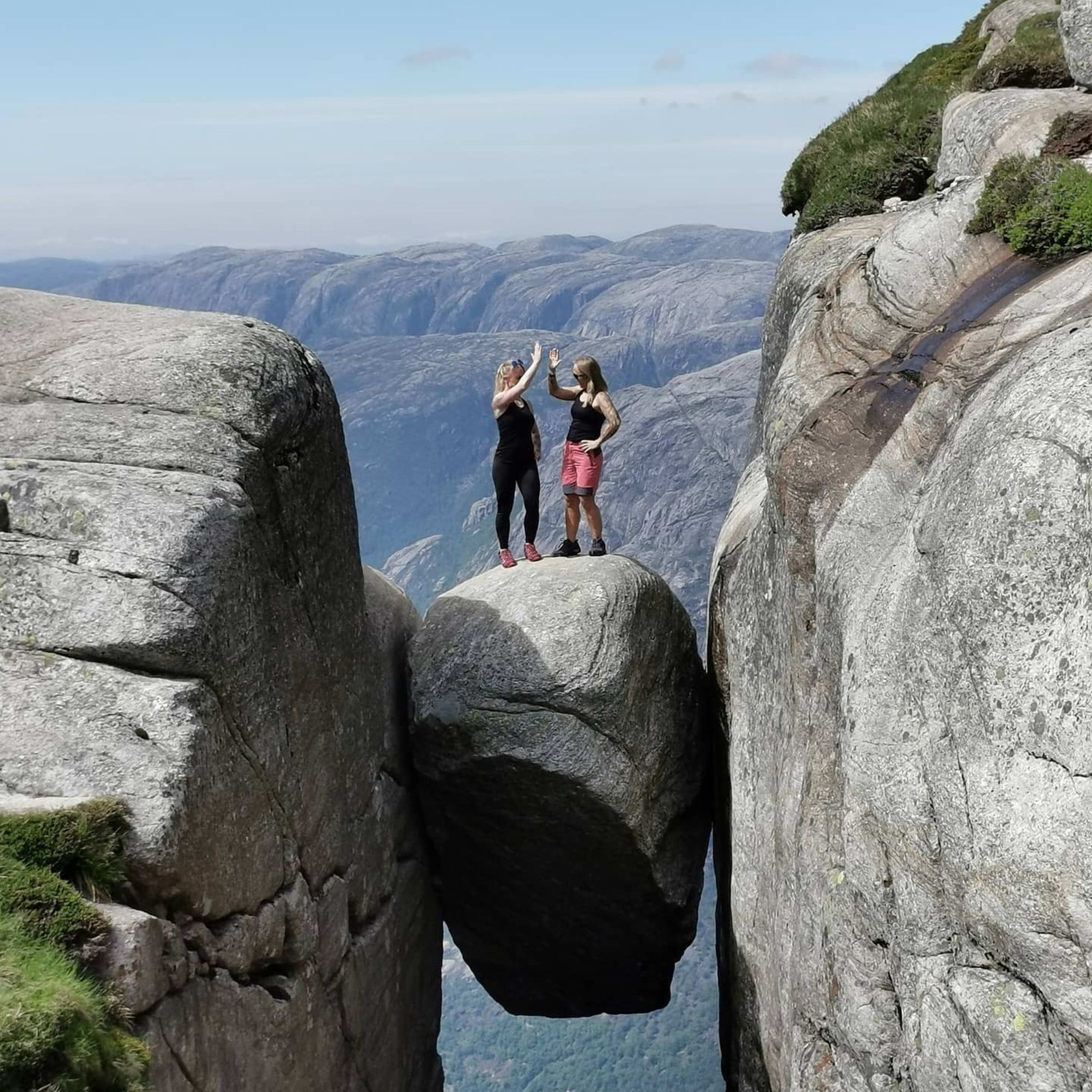 To kvinner gir hverandre high-five på Kjeragbolten, en stor stein som sitter mellom to massive fjellvegger over en dyp fjelldal.