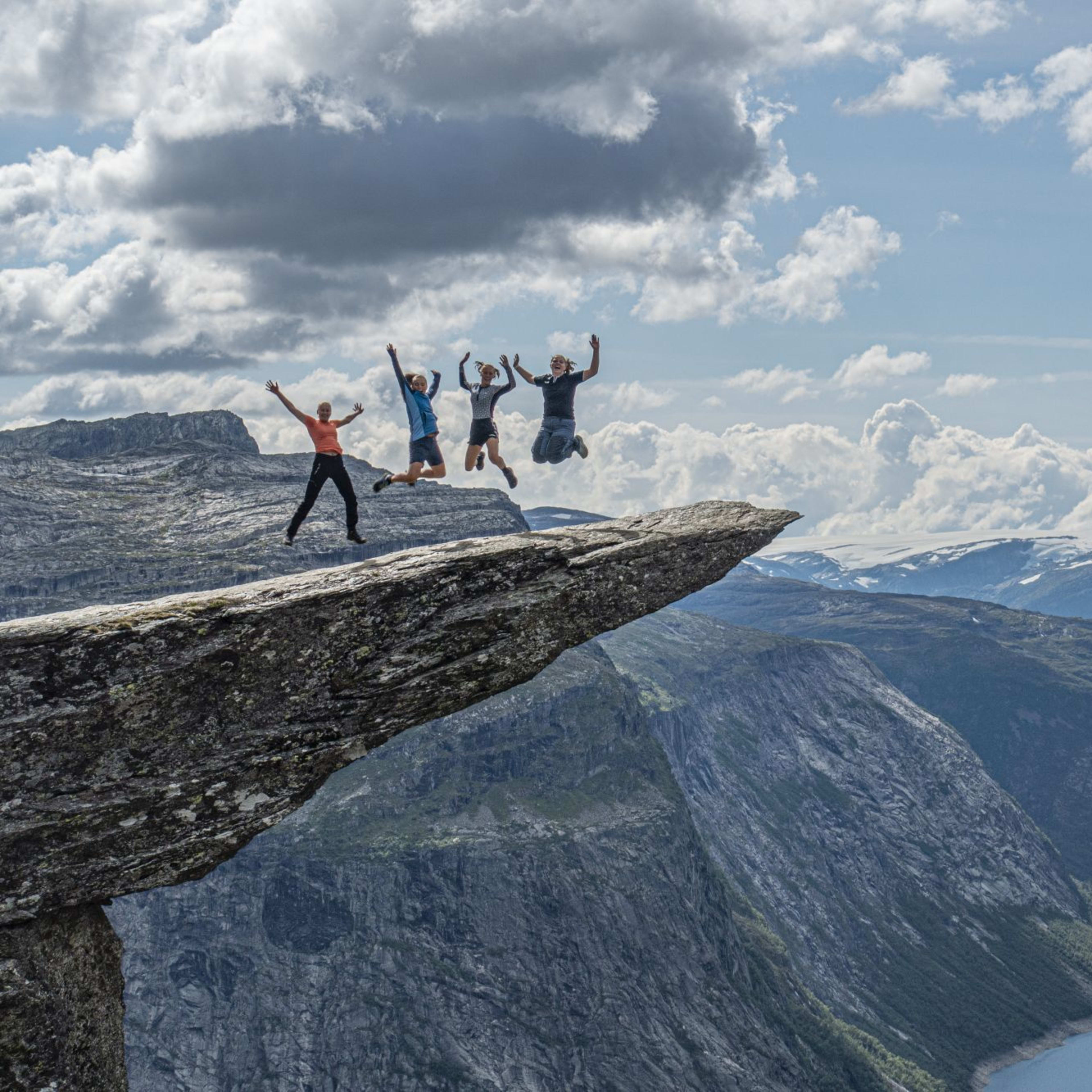Four people jump on Trolltunga above dramatic fjord and cliffs.
