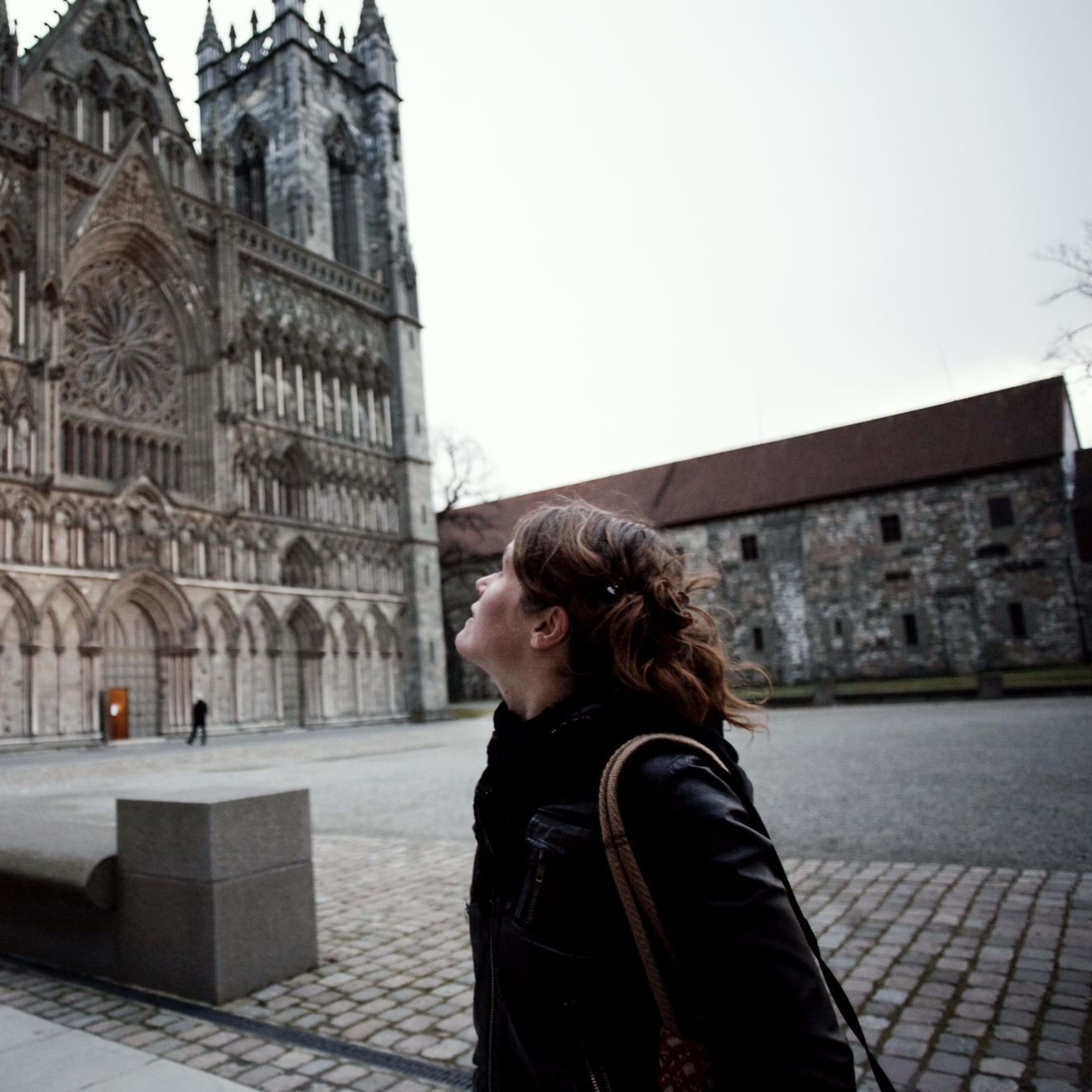Frau, die die kunstvolle Fassade der Nidaros-Kathedrale mit detaillierten Schnitzereien und Statuen betrachtet, unter einem bewölkten Himmel.