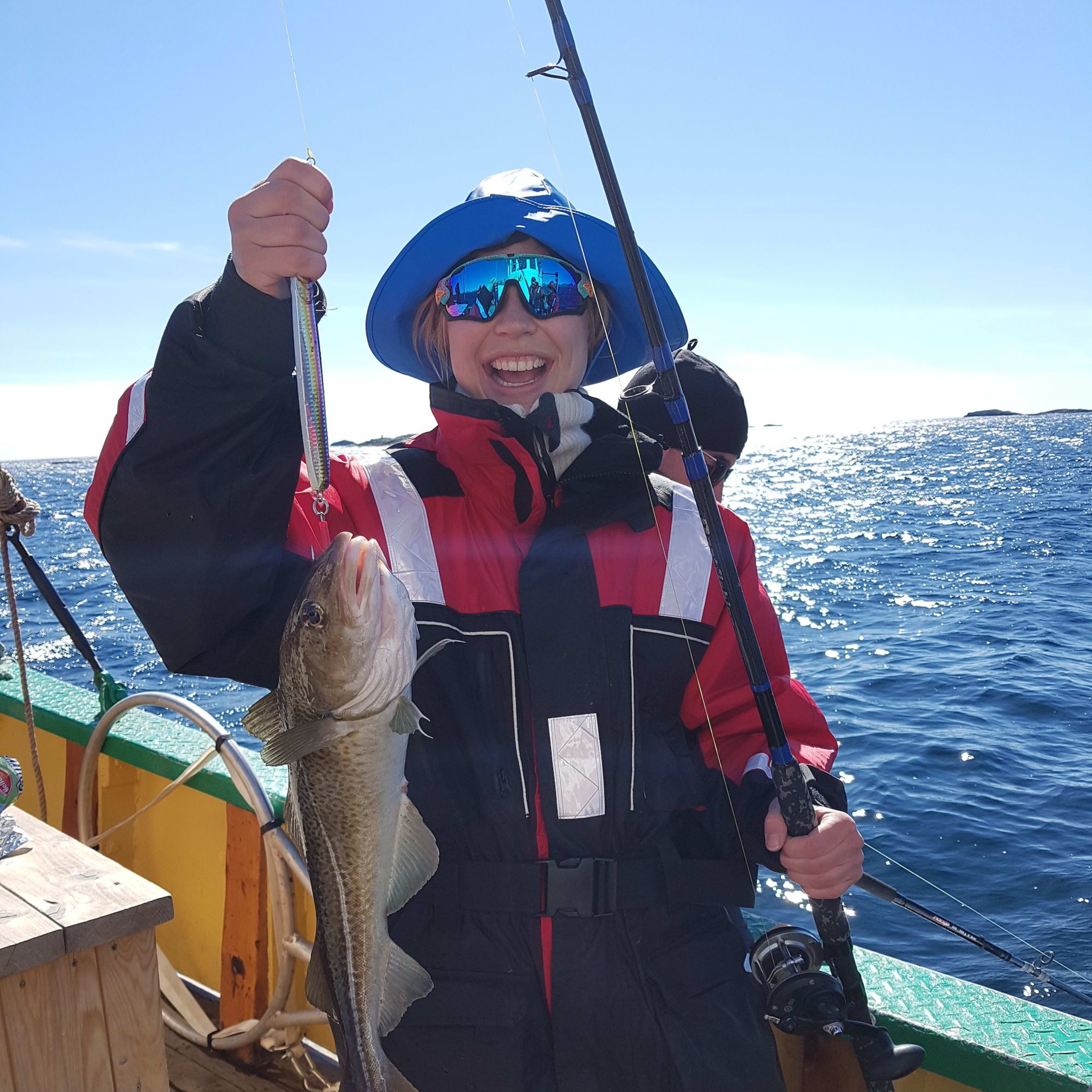 A woman holds up a freshly caught fish, smiling, on a sunny fishing day with calm waves.