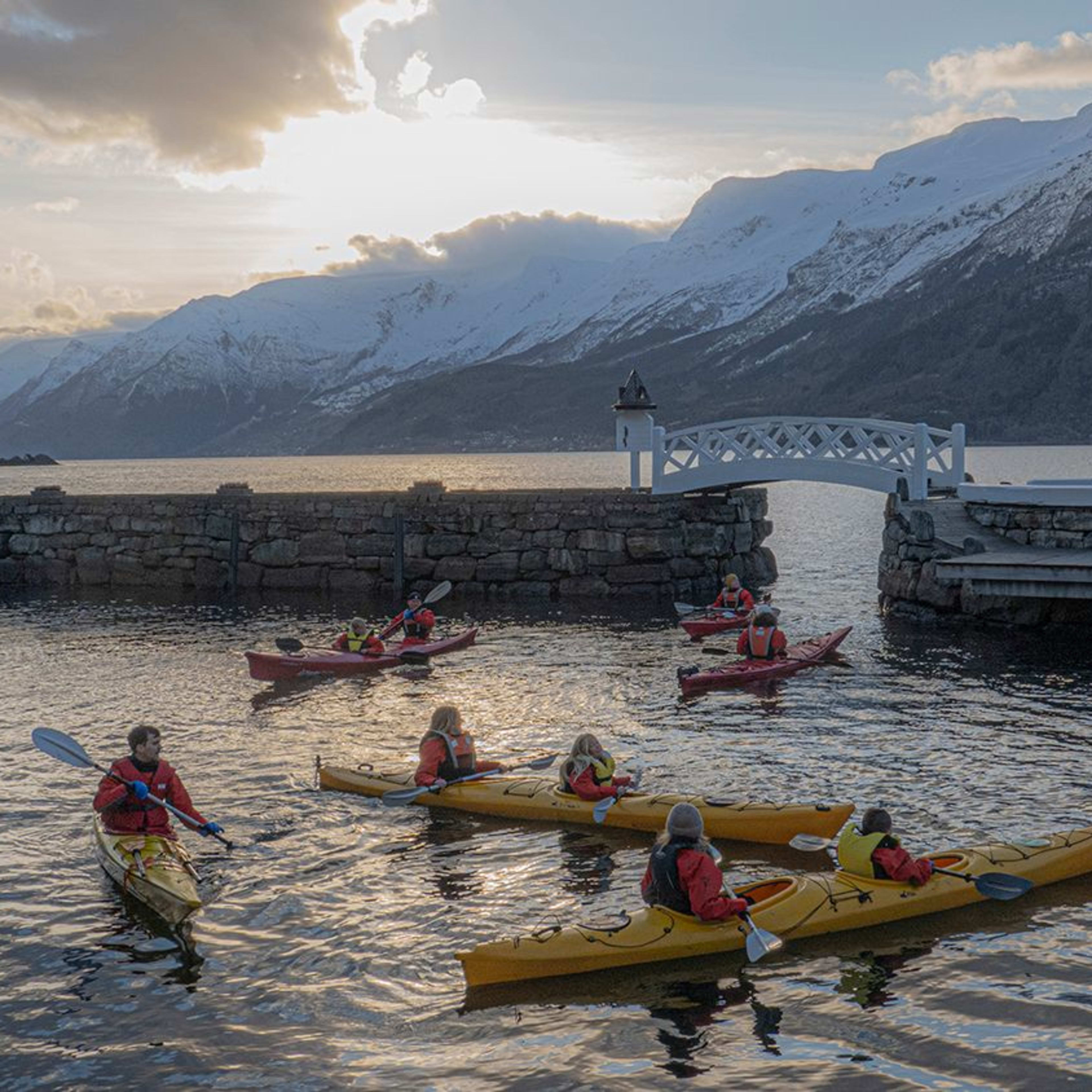 Eine Gruppe von Kajakfahrern paddelt auf ruhigem Wasser, mit einem Steinpier und schneebedeckten Bergen im Hintergrund.