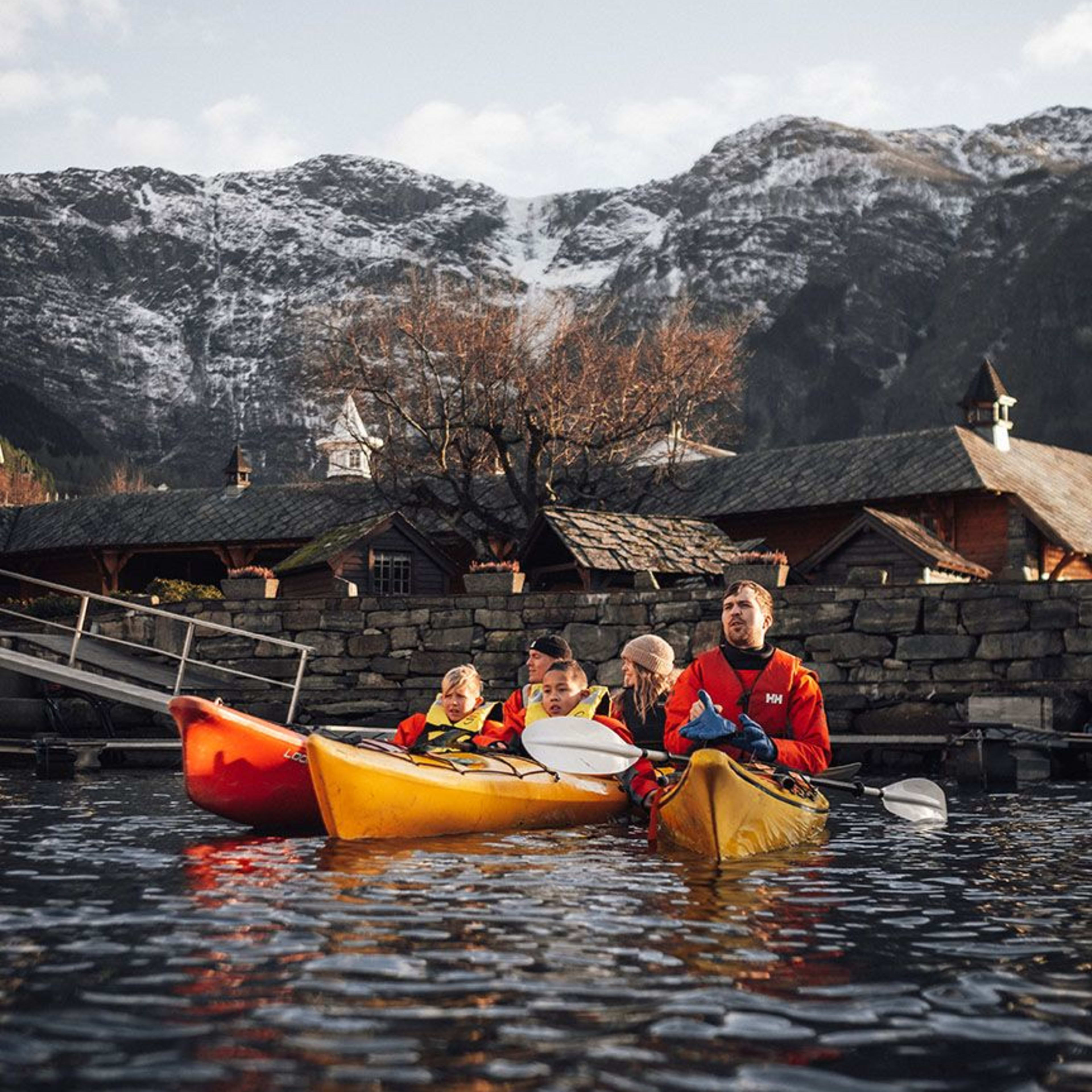 Eine Gruppe von Kajakfahrern paddelt nahe einer felsigen Küste mit schneebedeckten Bergen im Hintergrund.