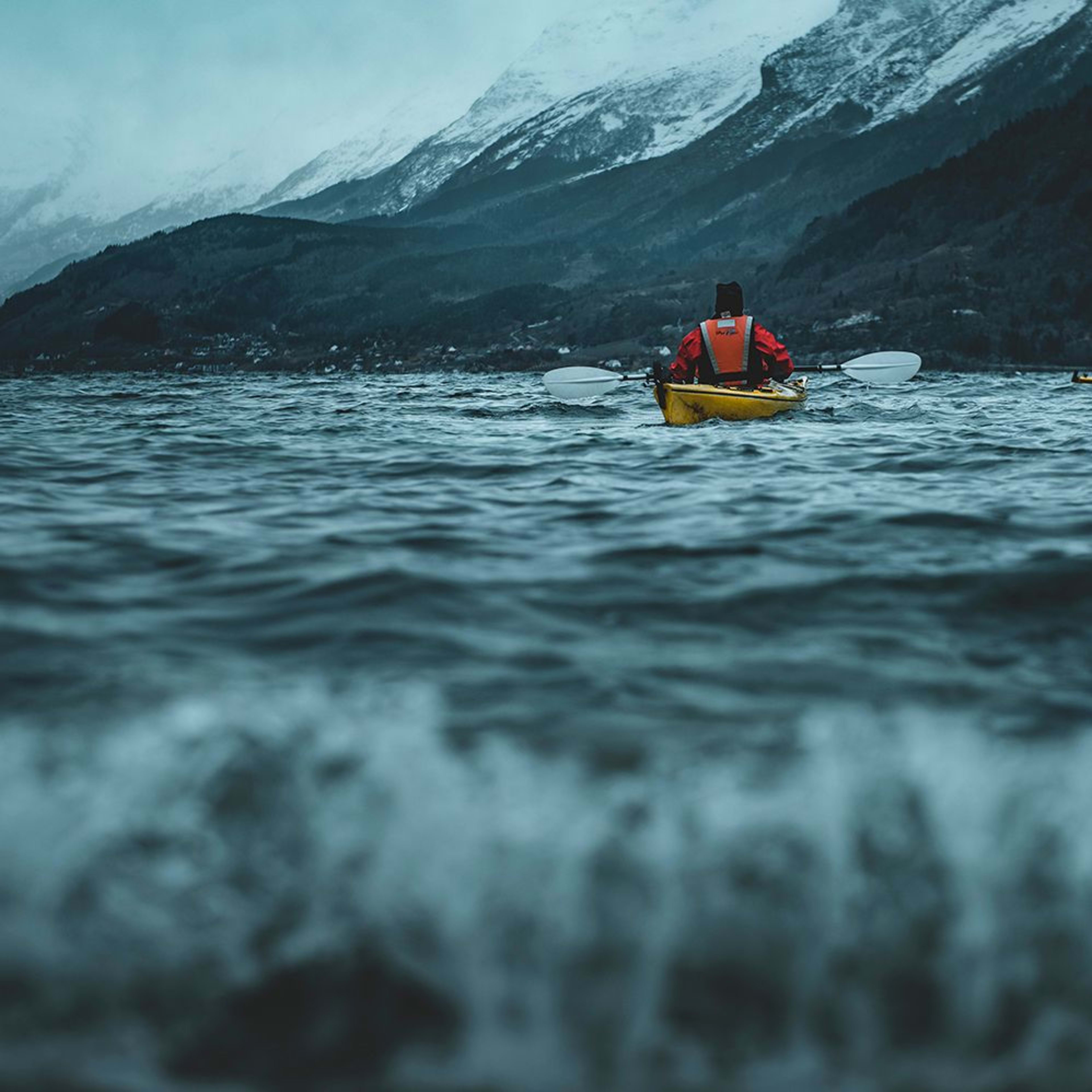 Zwei Kajakfahrer navigieren auf stürmischem Wasser mit schneebedeckten Bergen und einer kalten, dramatischen Atmosphäre.