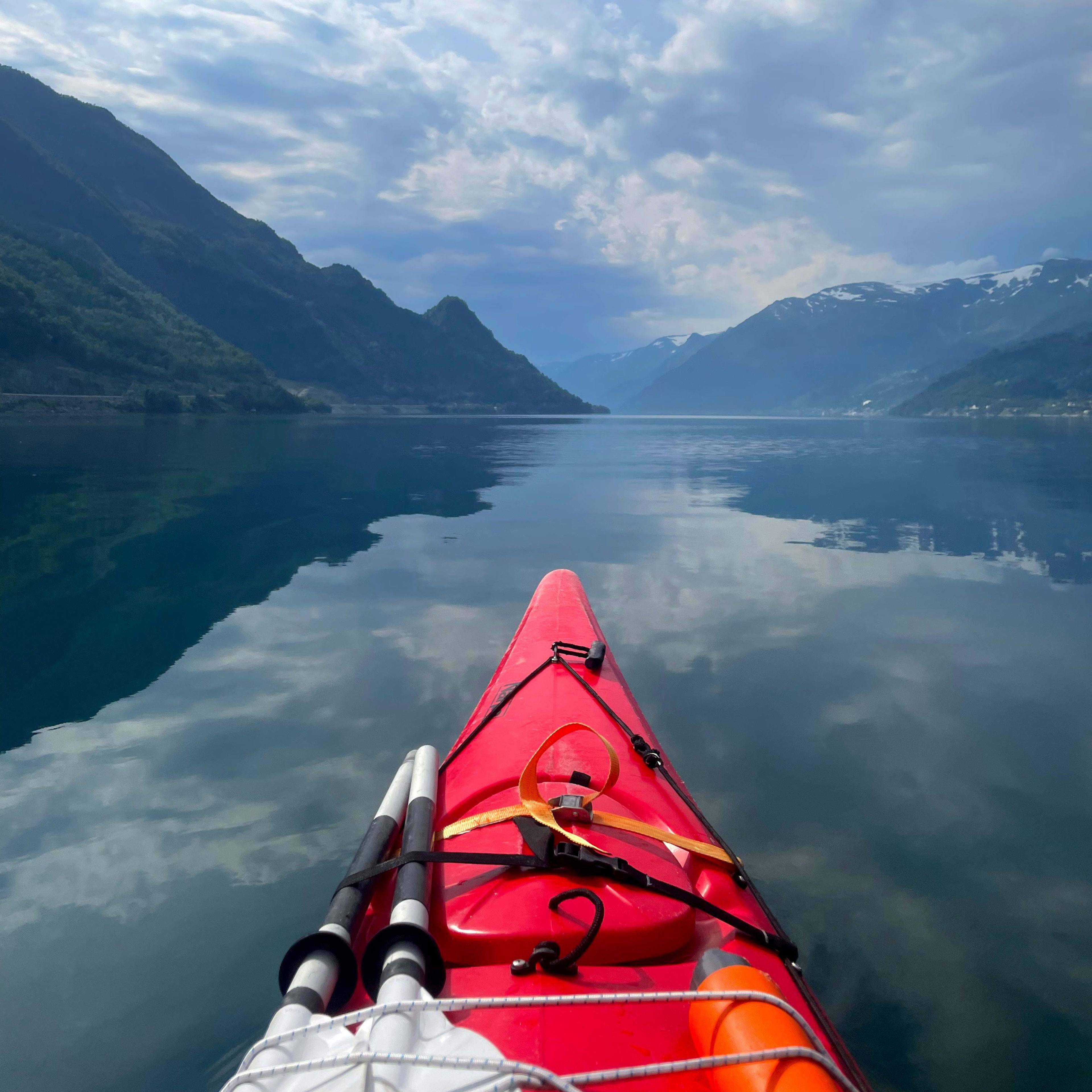 Eine friedliche Kajakszene mit einem roten Kajak, das ruhig durch Wasser schneidet und den Himmel und die Berge reflektiert.