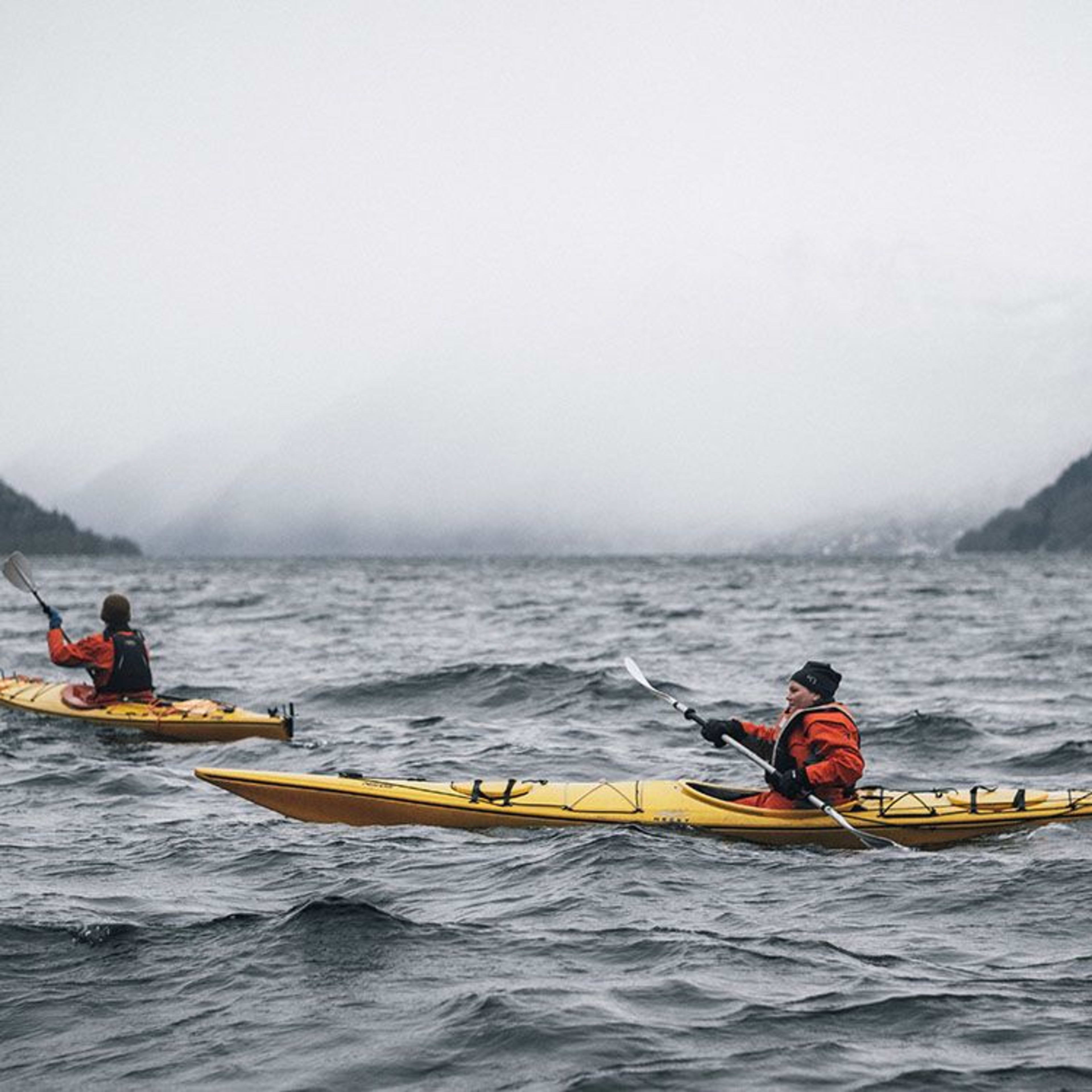 Kajakfahrer in gelben Kajaks auf stürmischem Wasser mit nebelverhangenen Bergen im Hintergrund.