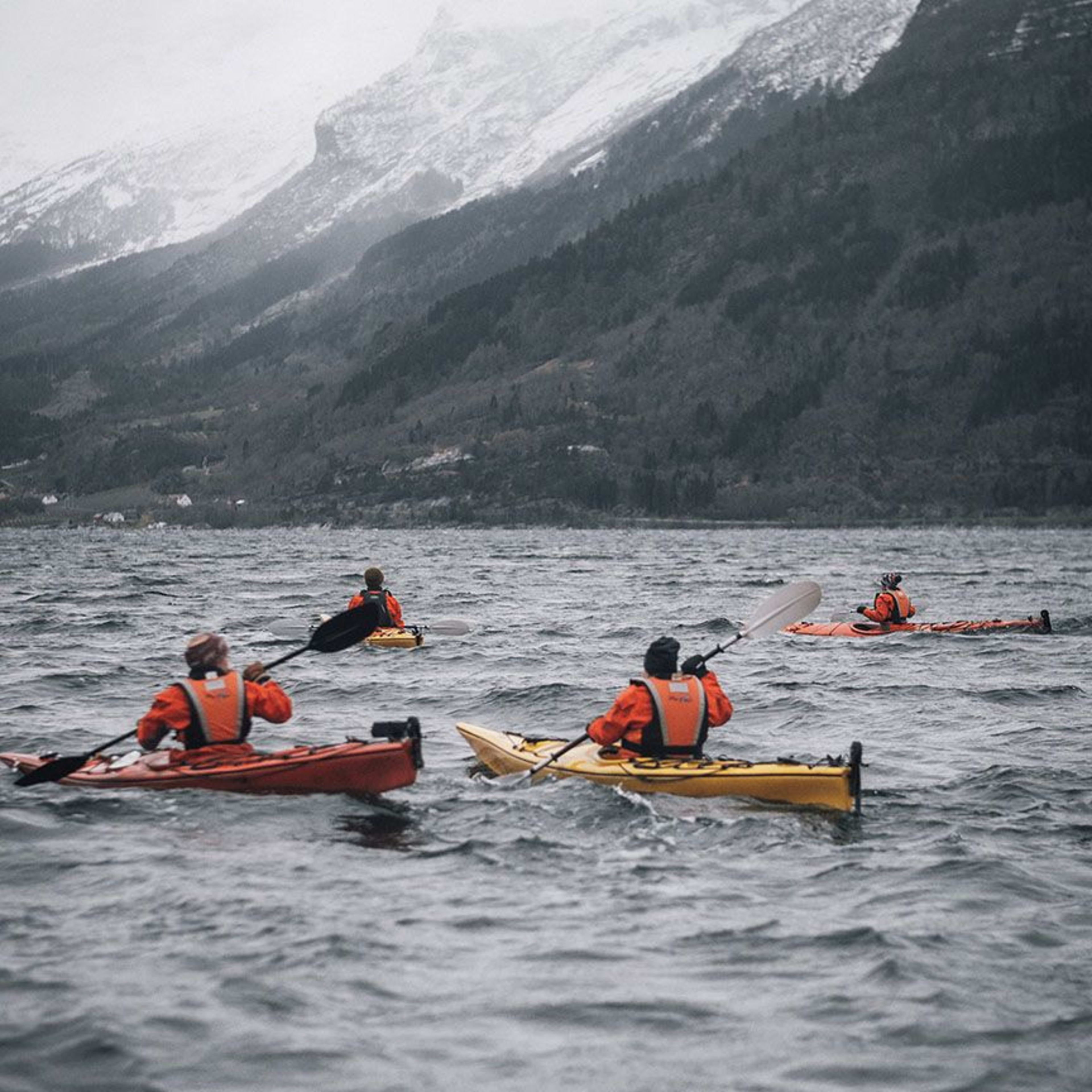 Kajakfahrer paddeln in stürmischem Wasser mit schneebedeckten Bergen im Hintergrund, was eine abenteuerliche Atmosphäre schafft.