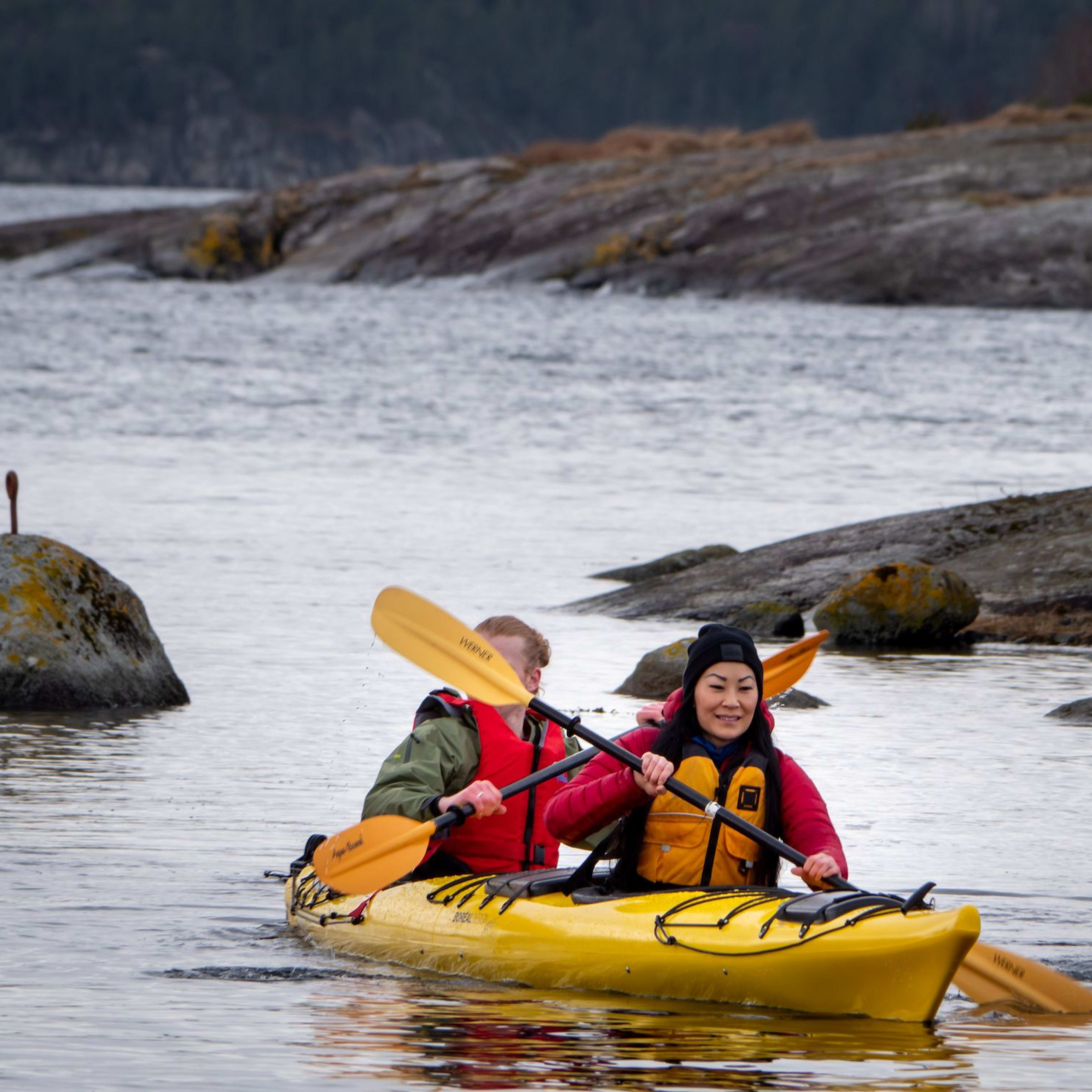 Two people paddle yellow tandem kayak through rocky coastal waters, both wearing life vests.