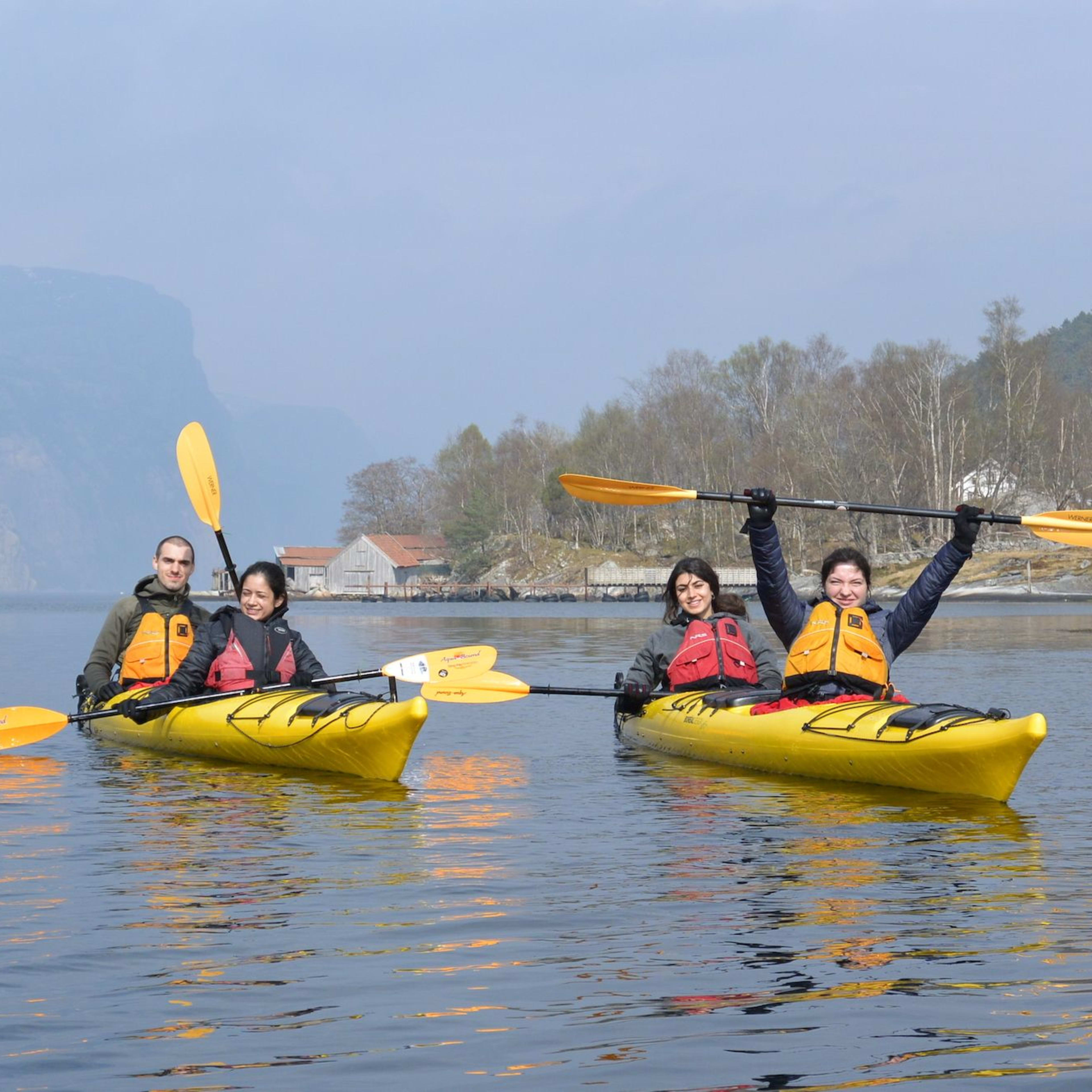 Four people paddle yellow kayaks on a calm fjord with misty hills.