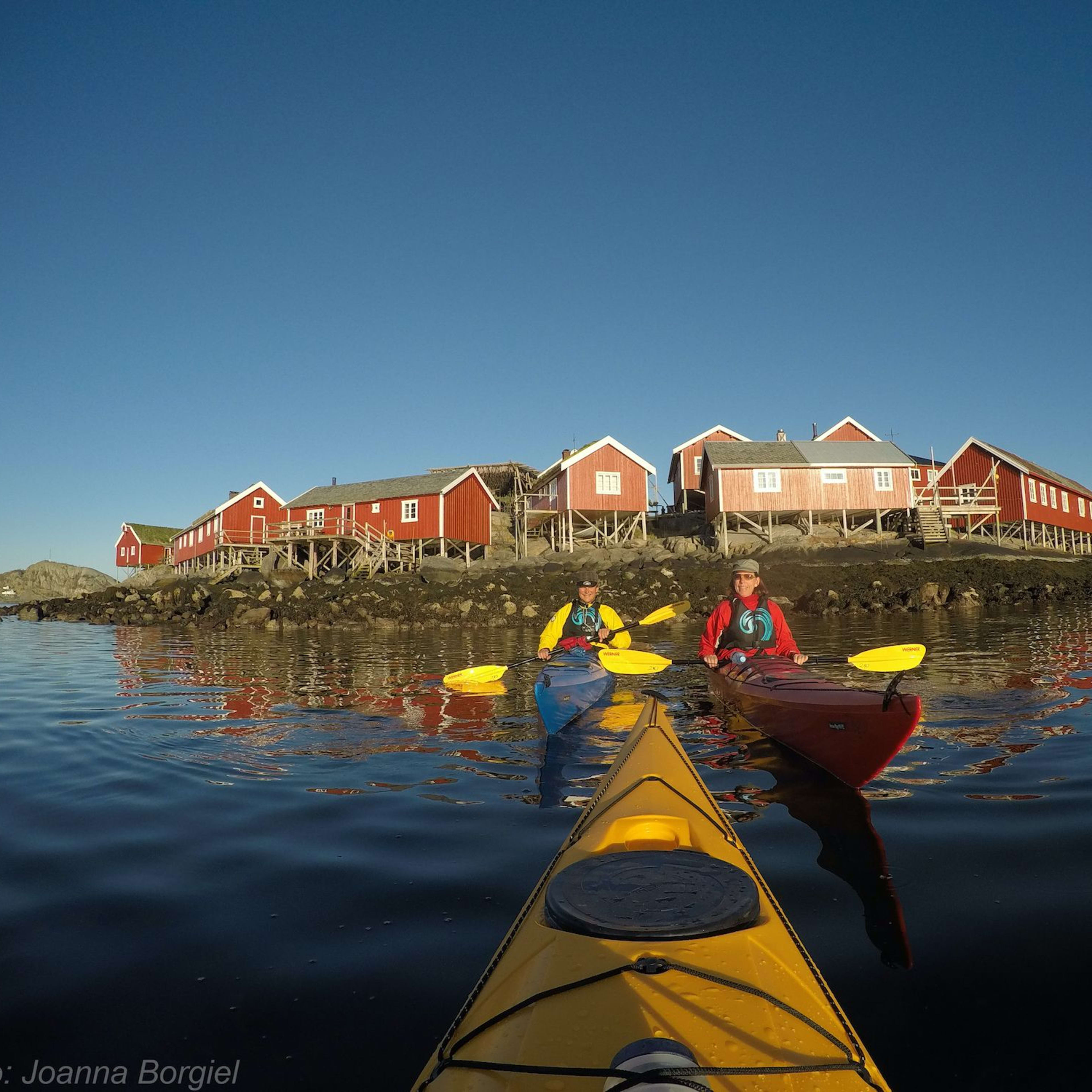 Two kayakers paddling under a clear sky with red wooden cabins on stilts lining the rocky shore and their reflections in the water.