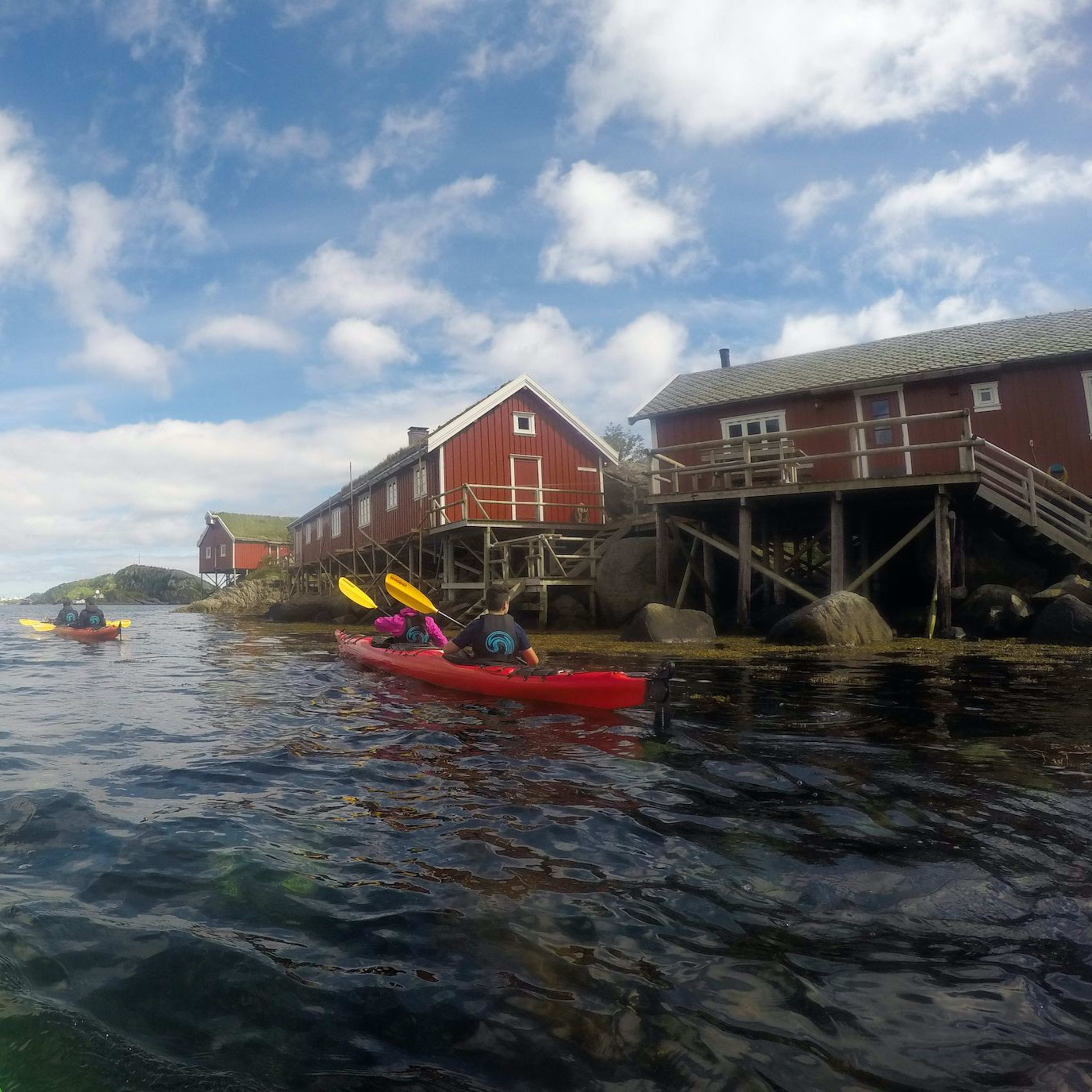 Colorful kayaks paddling by red wooden cabins on stilts, with clear skies and mountains in the distance.