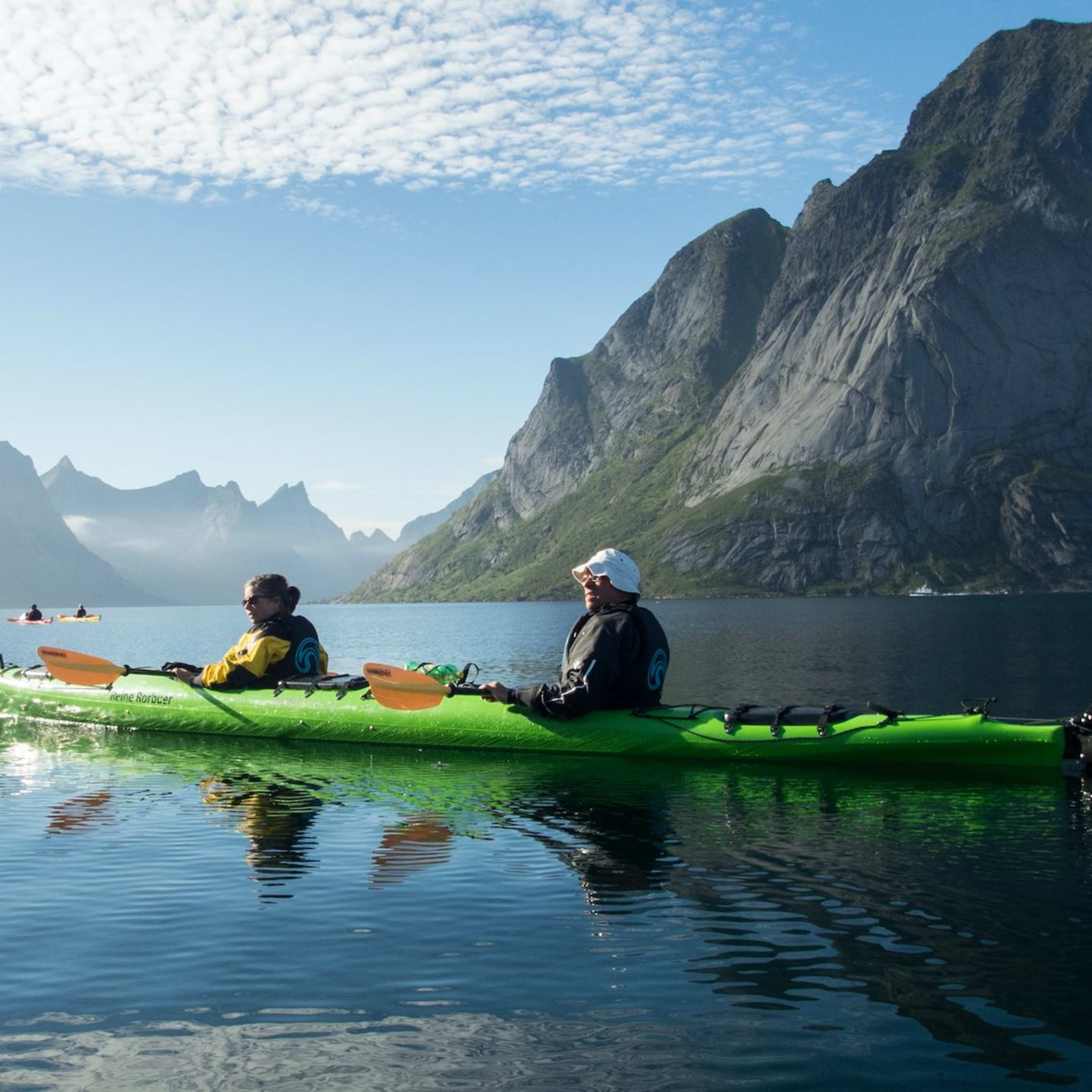Two kayakers paddling on calm water with towering mountains in the background, under a clear sky with wispy clouds.