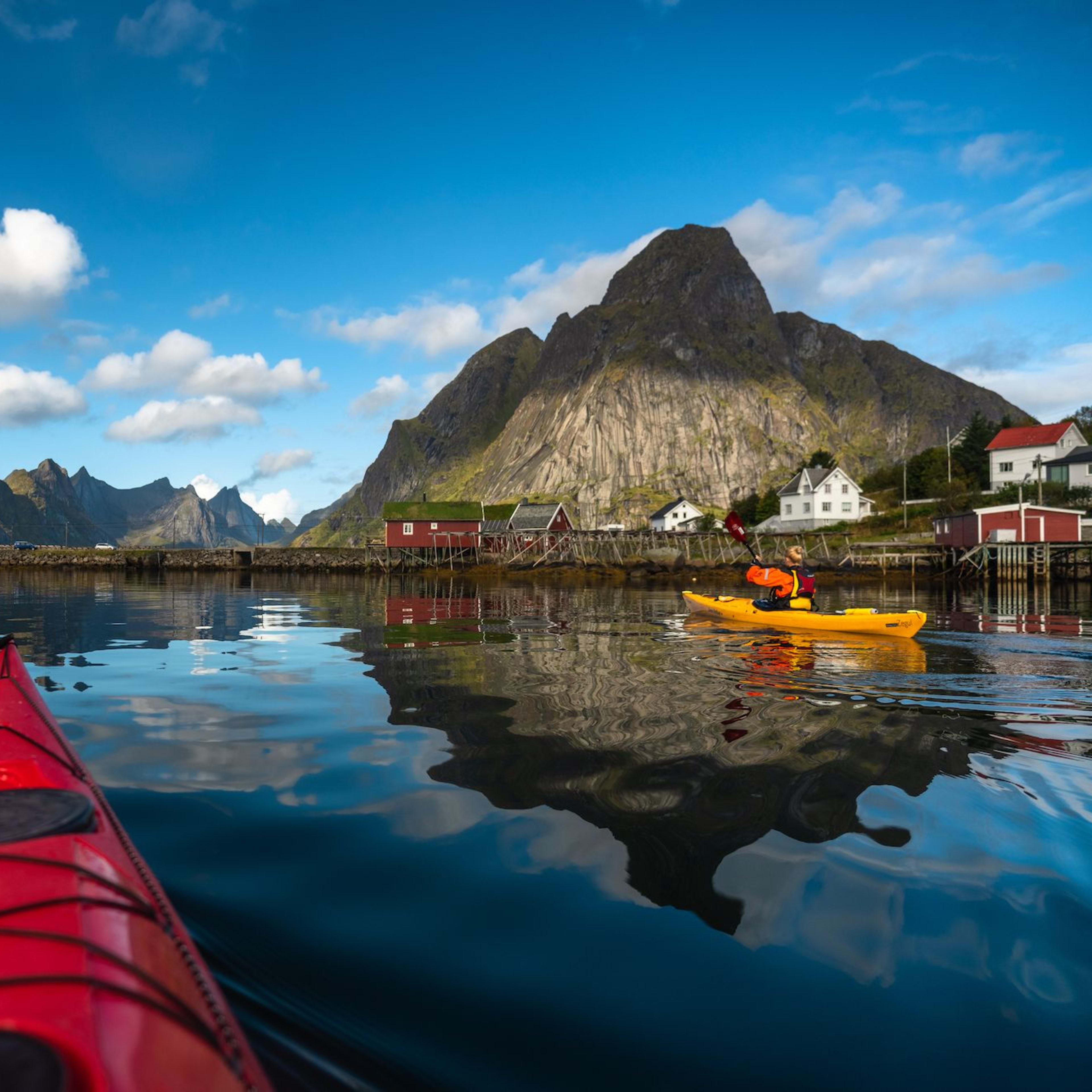 Kayakers paddling through calm waters with colorful houses on stilts and jagged mountains in the background.