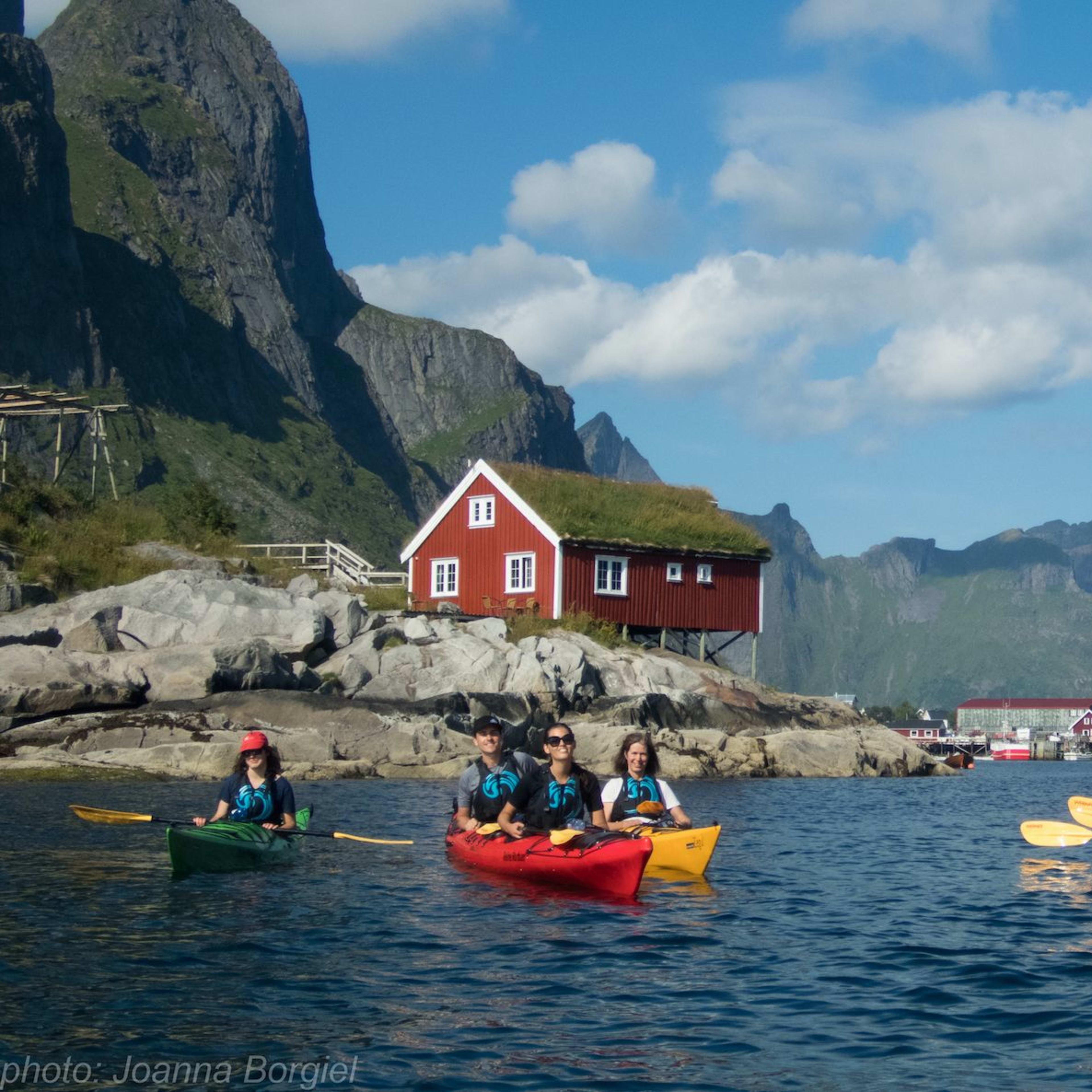 Kayakers paddling near a small island with a red house, green mountains, and blue sky, creating a peaceful and adventurous scene.