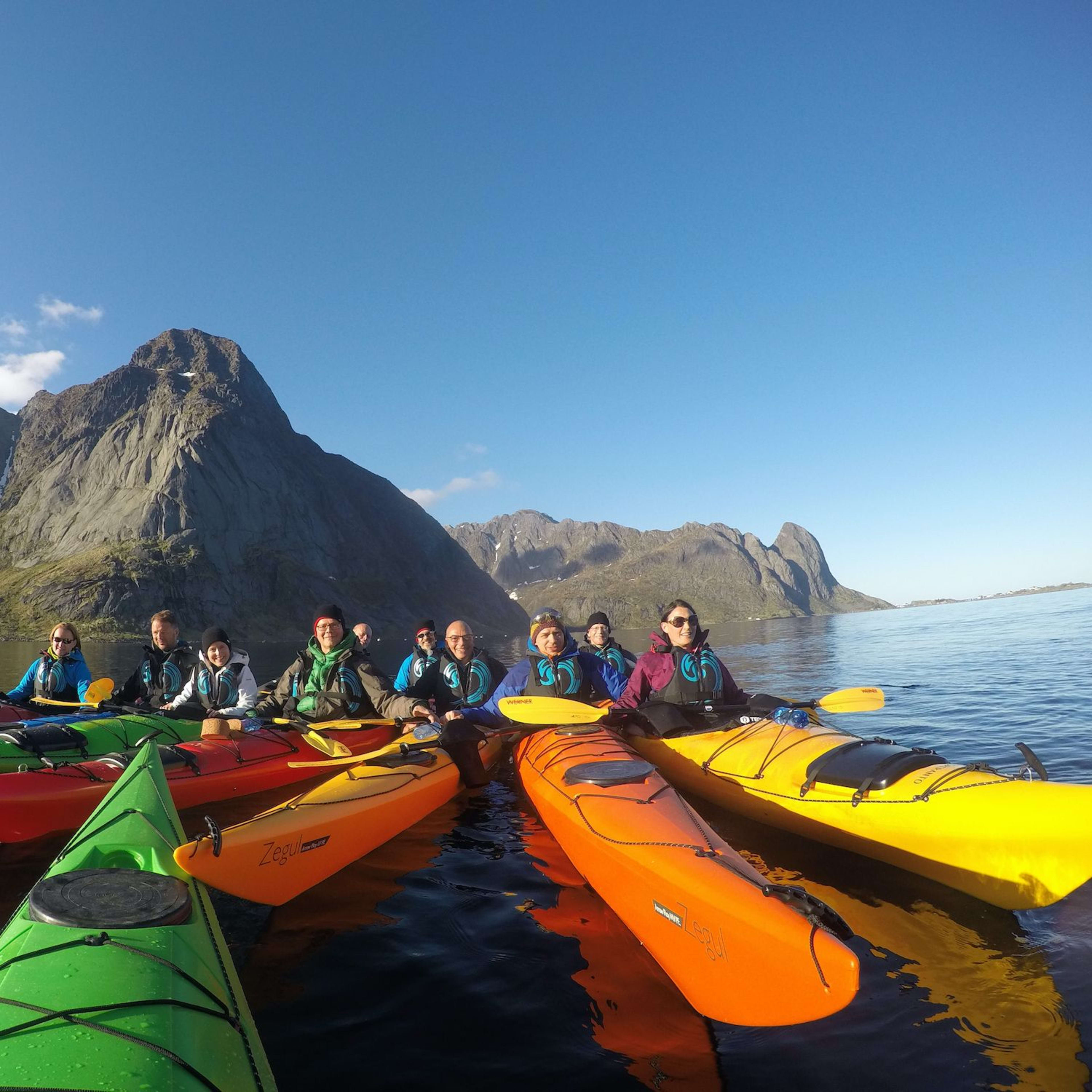 Kayakers gathered in colorful kayaks, smiling on calm, clear water with dramatic rocky mountains in the background.